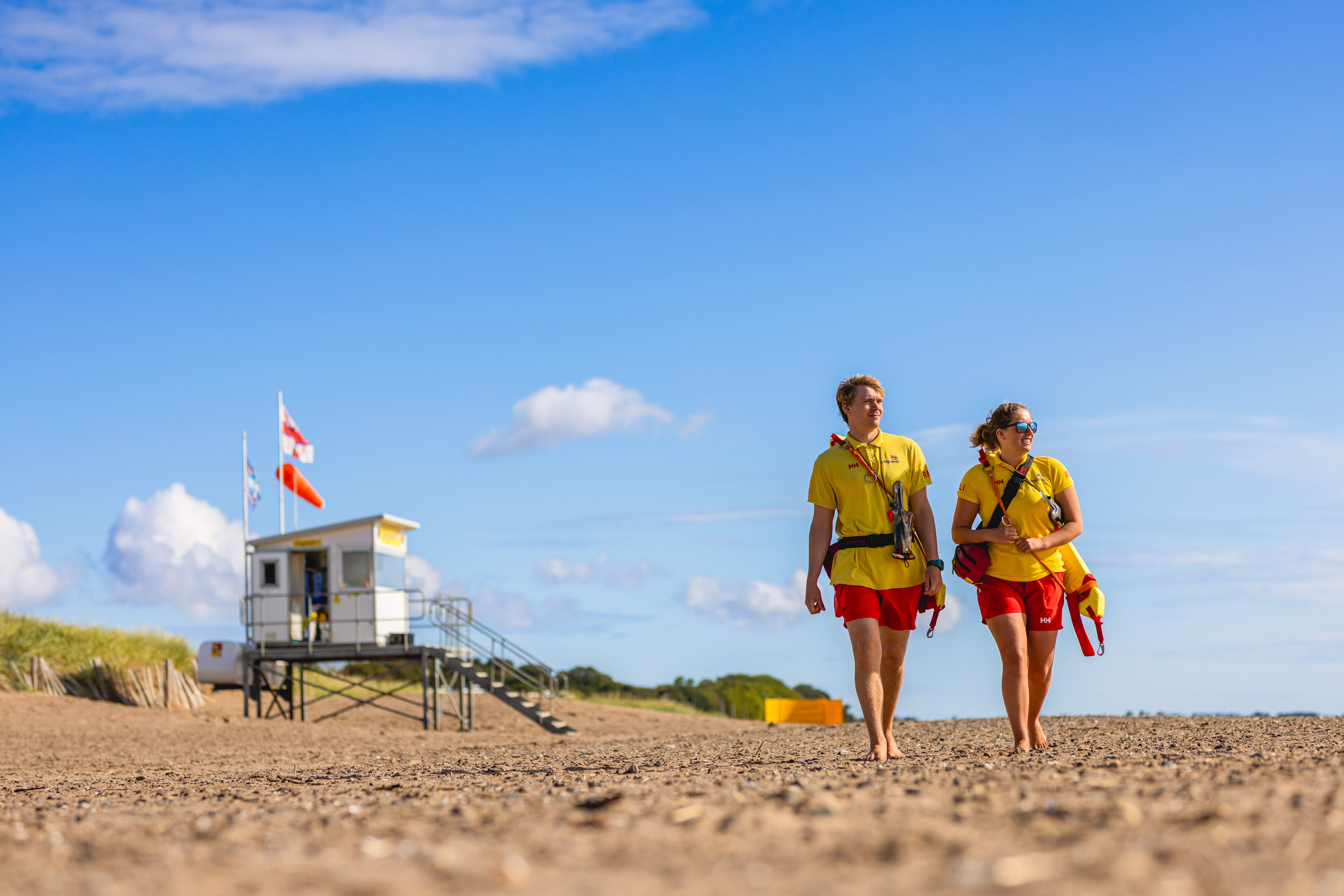 A female and a male RNLI lifeguard stroll across a beach with their lifeguarding hut in the background