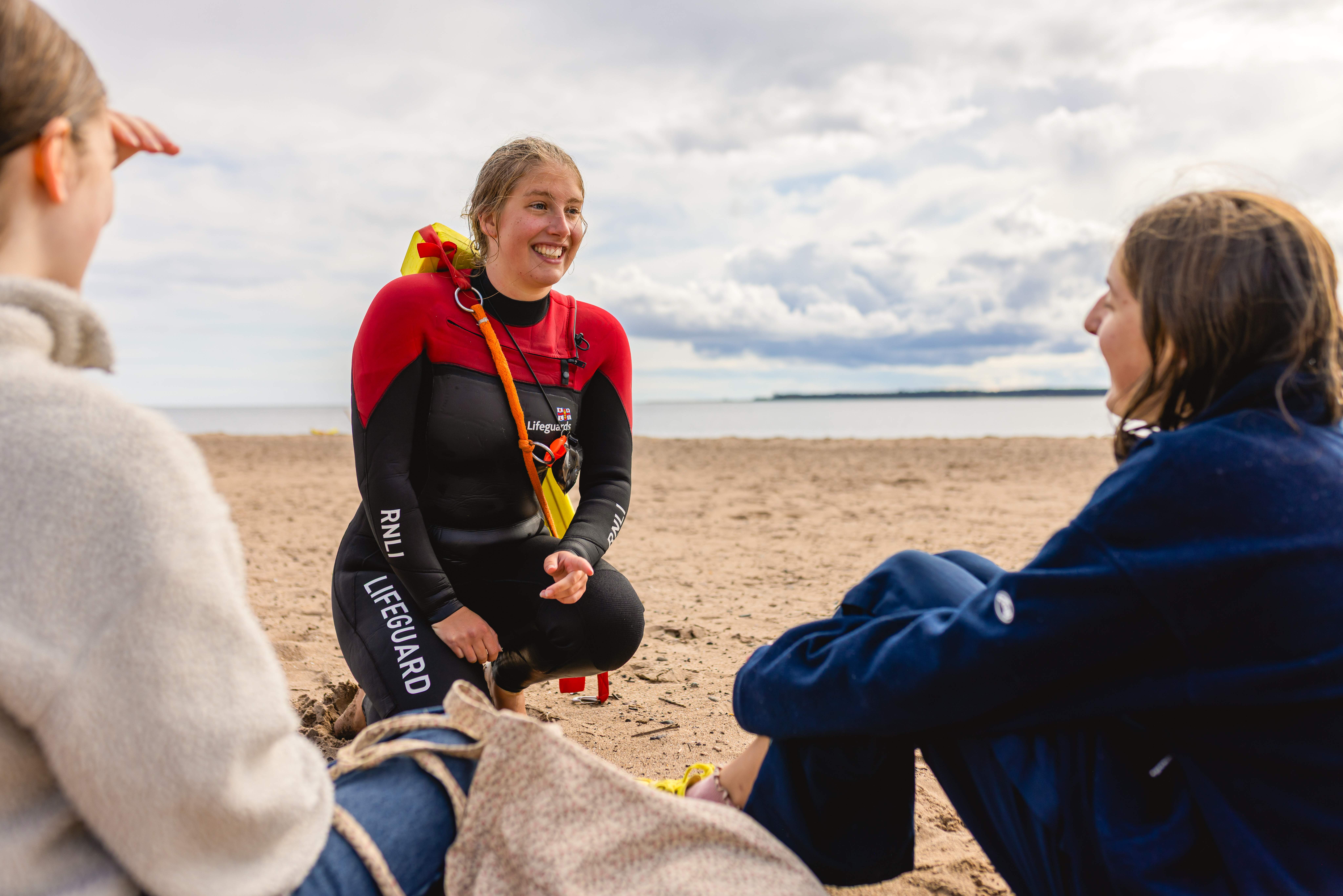 An RNLI lifeguards talks to two members of the public on a beach.