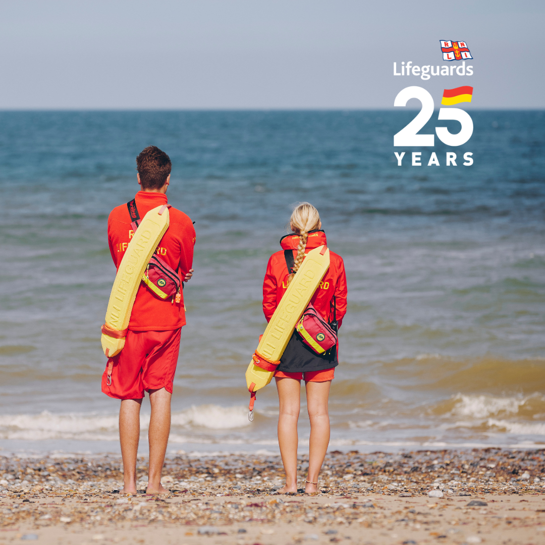 A female and male lifeguard stand on a beach and look out towards the sea