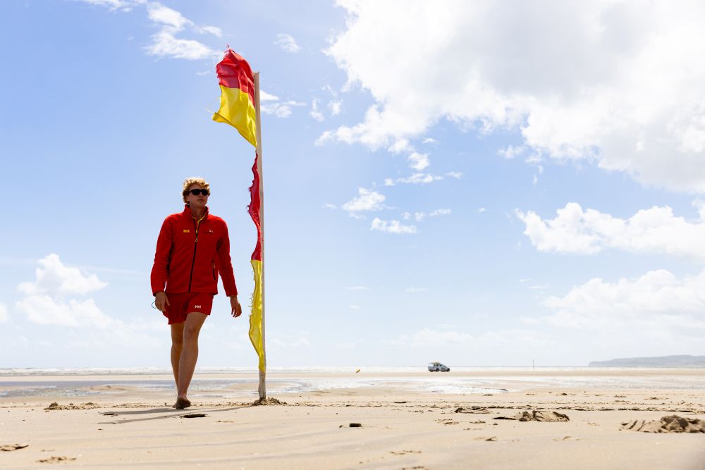 RNLI lifeguard walking along the beach next to a red and yellow flag