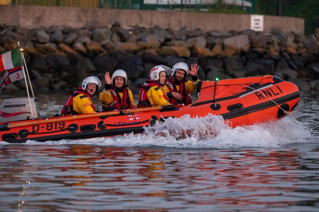 Four Fethard RNLI crew members dressed in yellow drysuits and red lifejackets in an orange D-class lifeboat. 2 crew members are waving