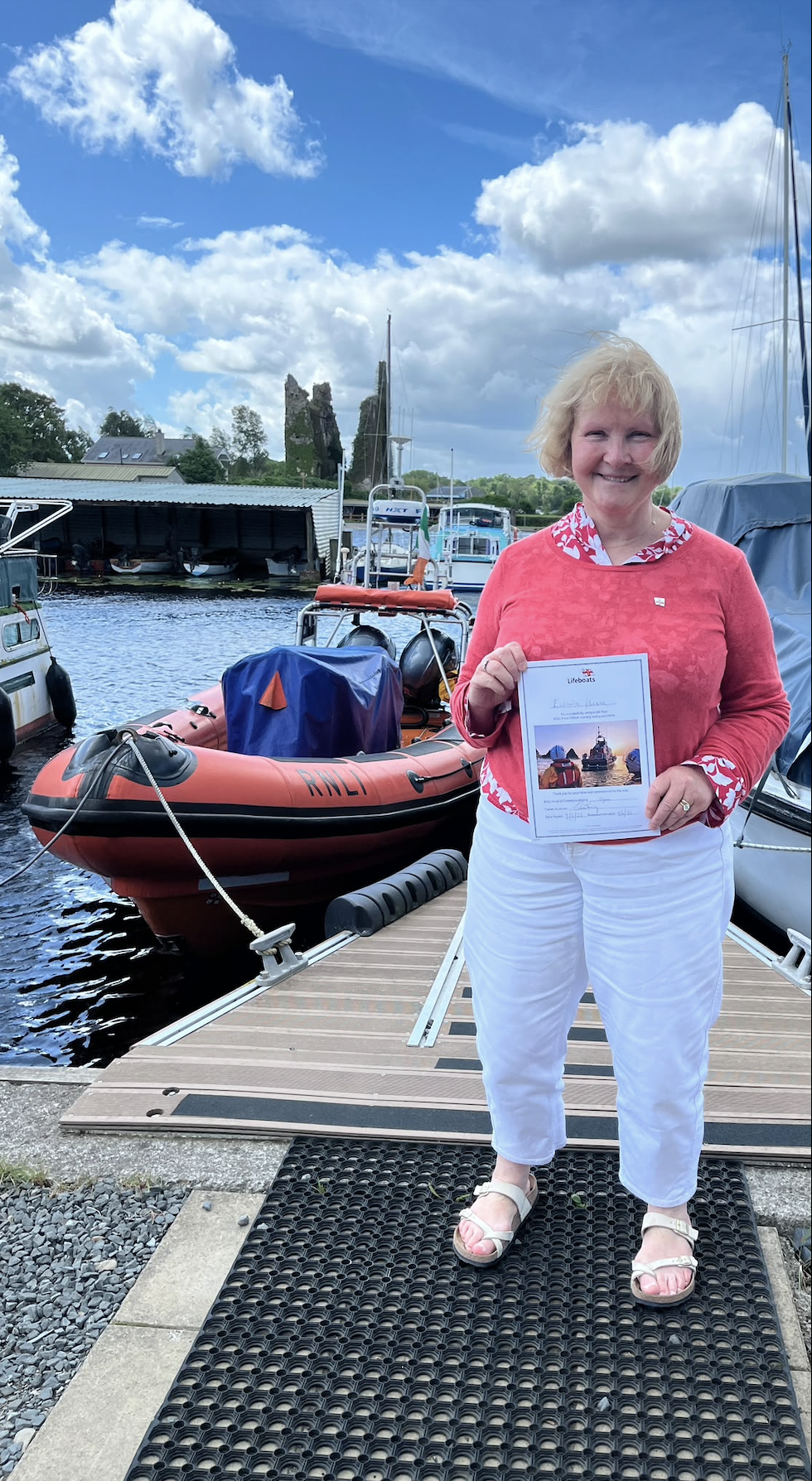 Eleanor stands on shore in front of the lifeboat holding her Lifeboat Press Officer assessment certificate