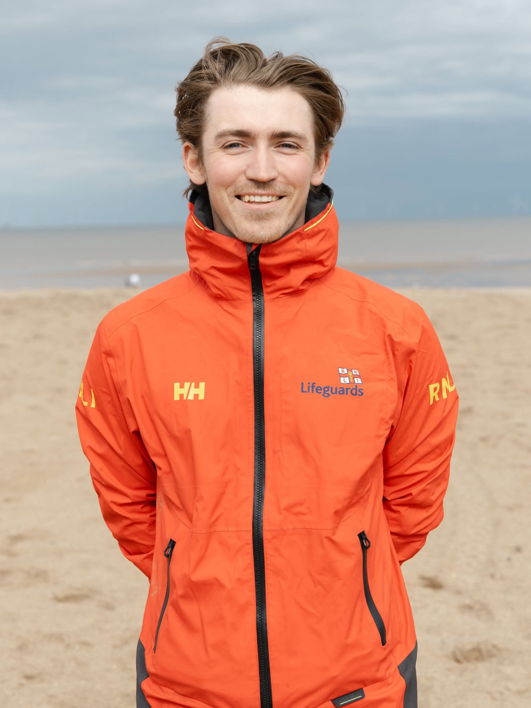 A male RNLI lifeguard stands for a photograph. He can be seen from the waist up while wearing a bright orange jacket. A sandy beach can be seen behind the RNLI lifeguard.