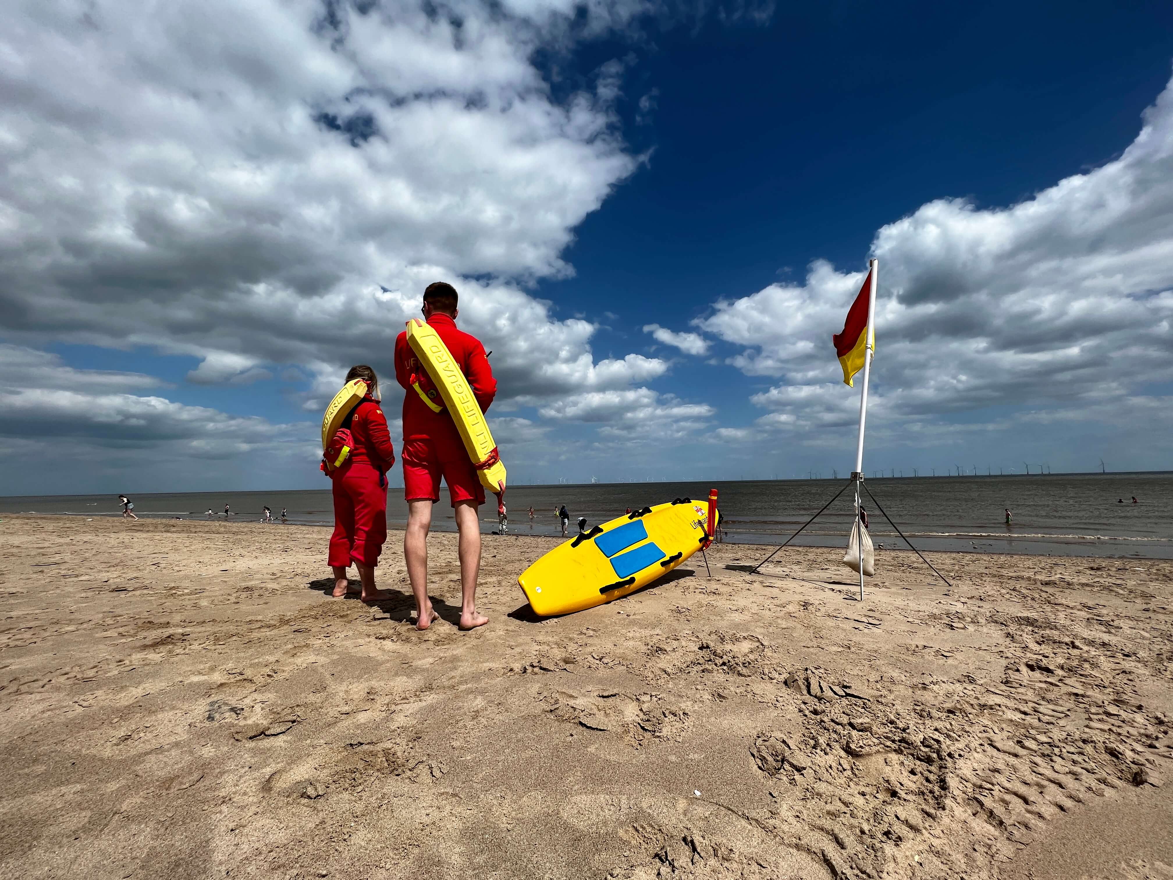 Two RNLI lifeguards stand upon the beach while on patrol. They have their backs turned to the camera. A yellow rescue board sits on the sand. A red and yellow lifeguarding flag blows in the wind nearby. Conditions are sunny and dry.