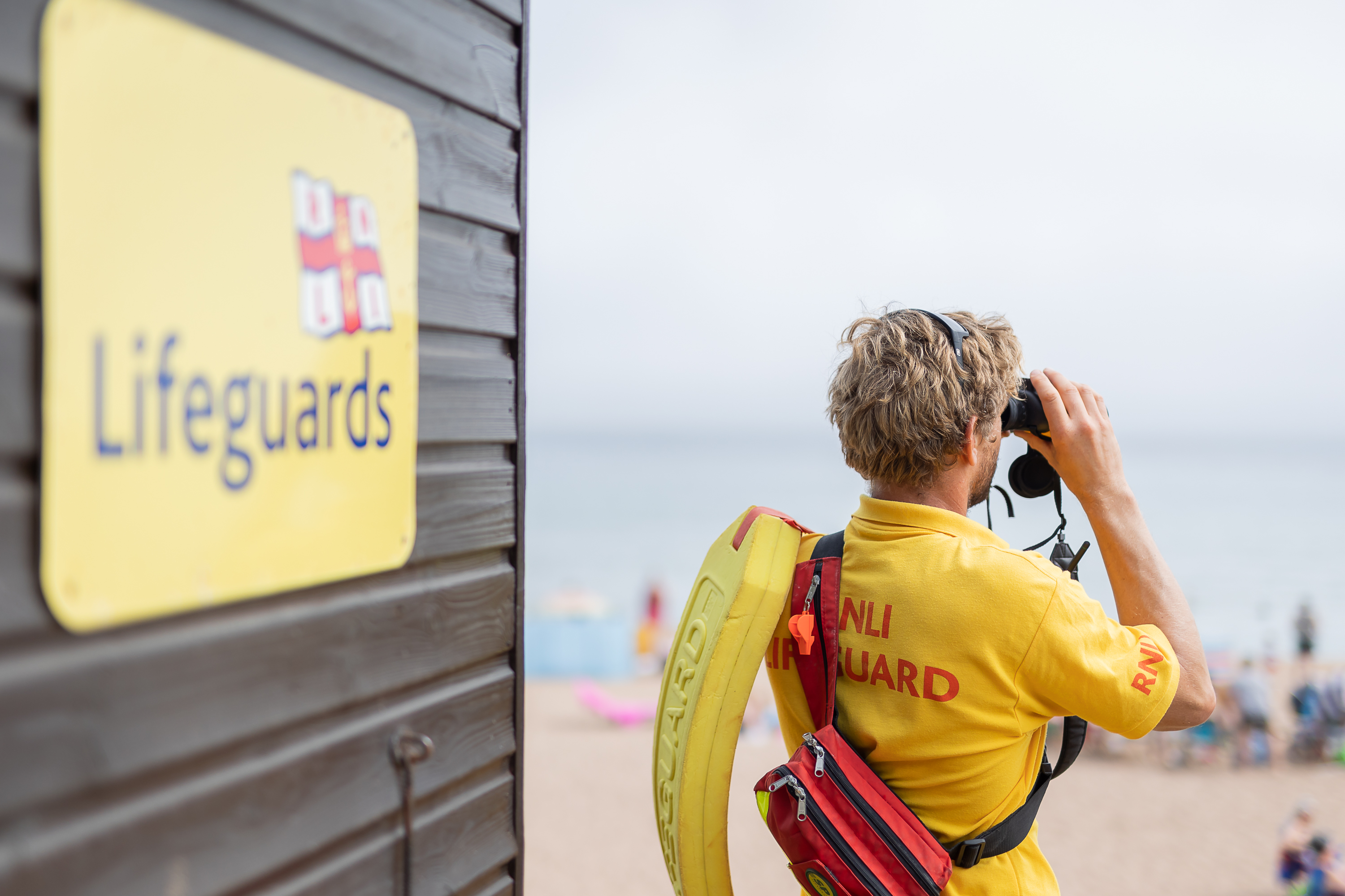 A RNLI lifeguard maintains a vigilant watch over beachgoers through binoculars from a nearby lifeguard unit.