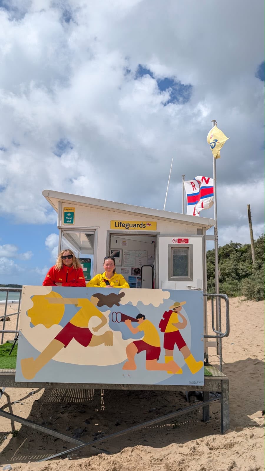 Portrait image of beach lifeguard unit with artwork in front of it. Two lifeguards stood next to it.
