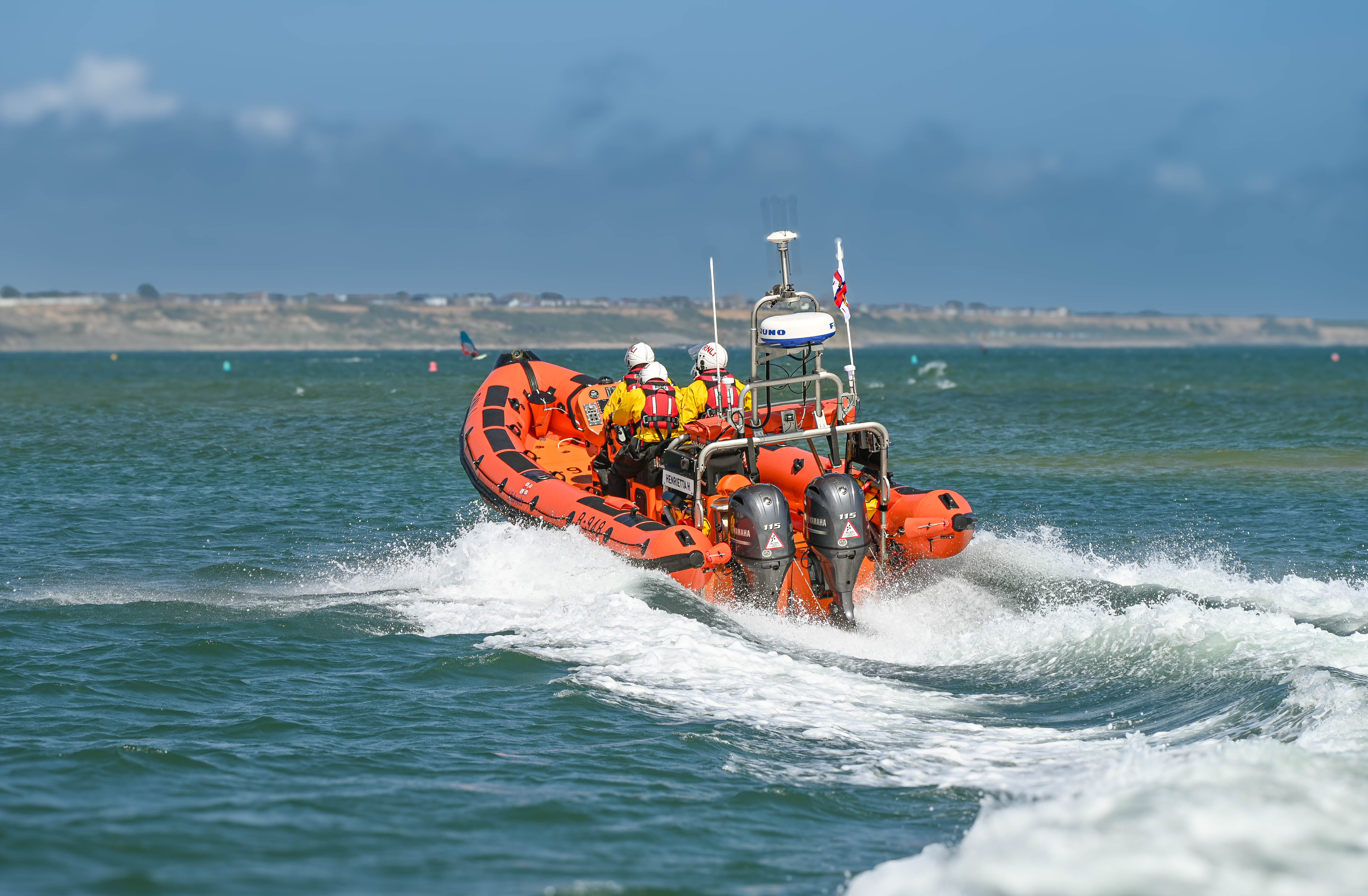 Four crew in yellow dry suits with white helmets and red lifejackets aboard an orange rib head out to sea.