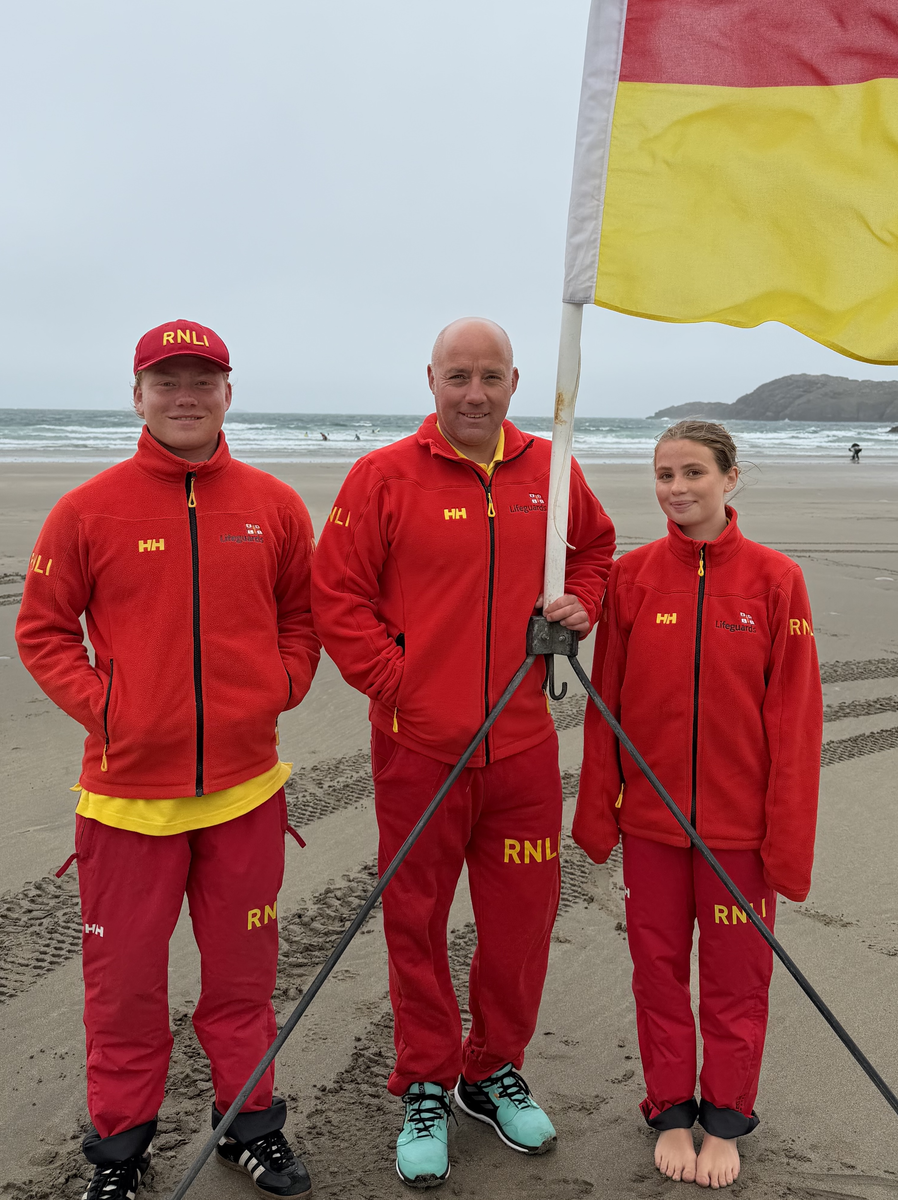 Portrait image of the Owen family stood in front of red and yellow flag on beach