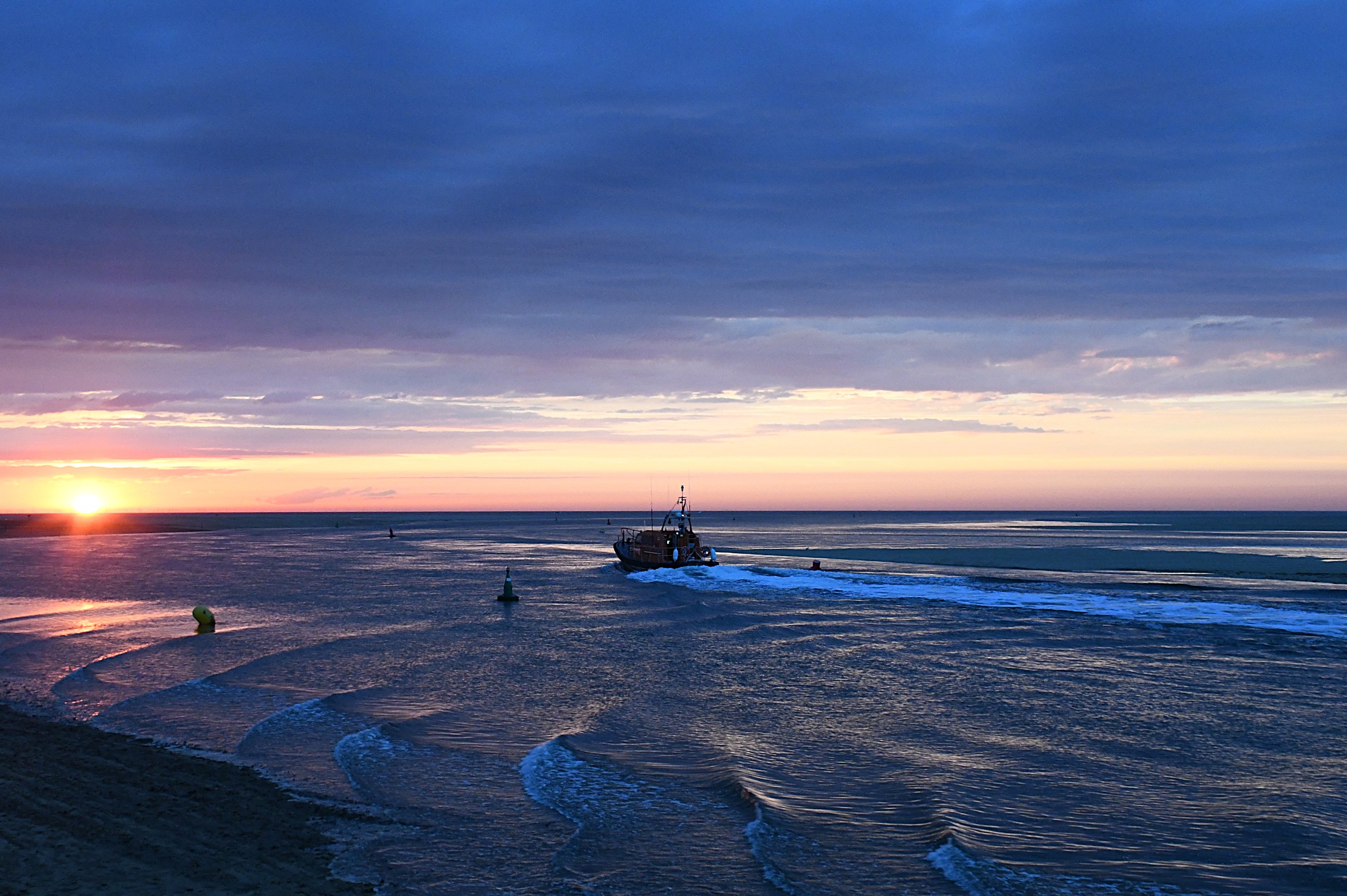 Wells RNLI Shannon class lifeboat on calm sea headed out of the harbour with an orange sunset in the background