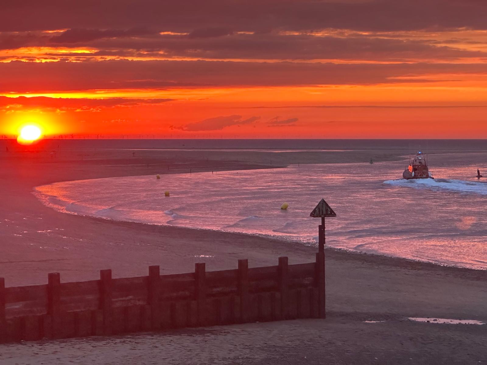 Wells RNLI Shannon class lifeboat on calm sea headed out of the harbour with an orange sunset in the background