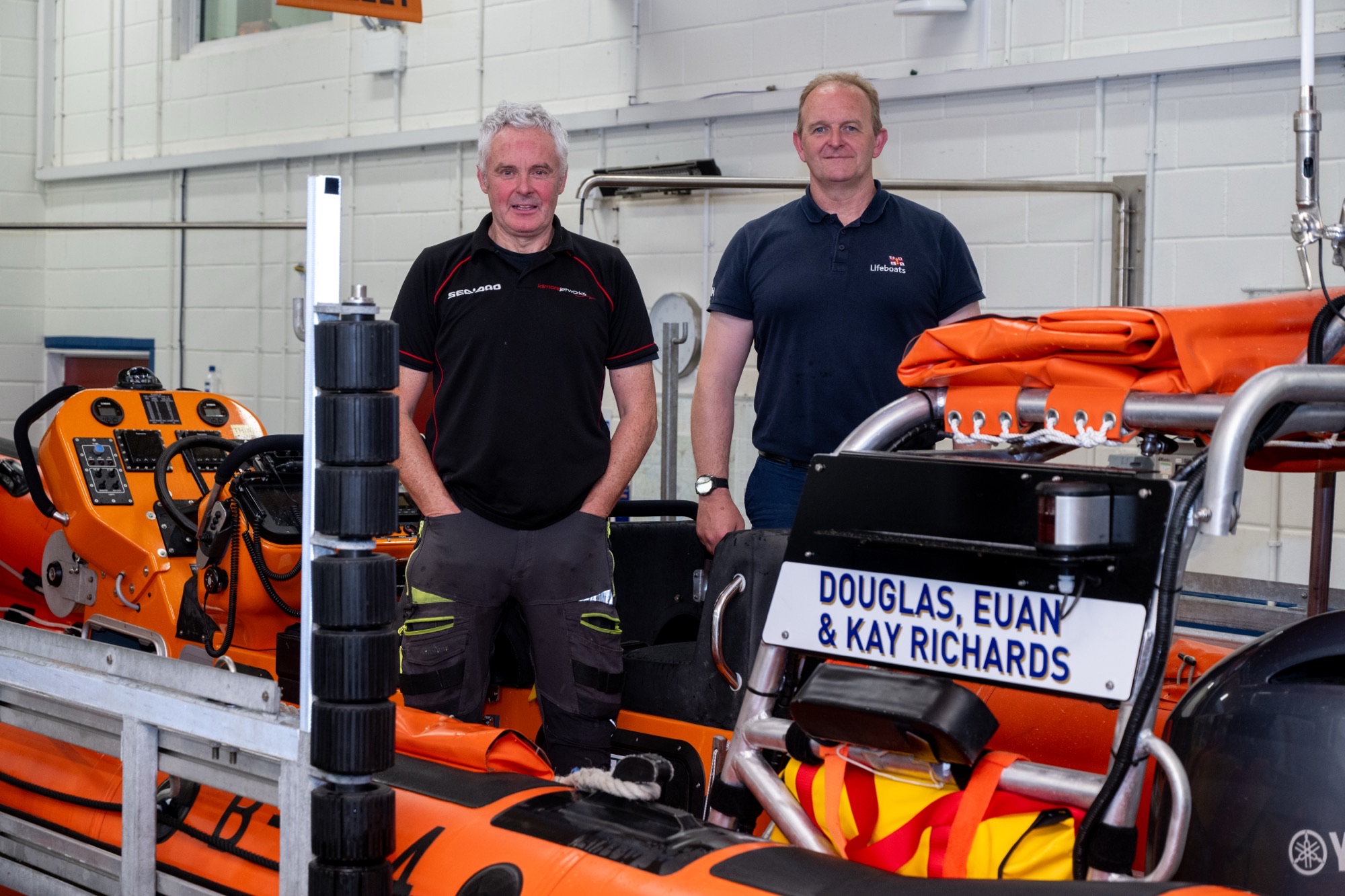 On the left of the picture is Carrybridge RNLI Helm, David Reid and on the right is Coastal Lifeboat Trainer, Sean Ginnelly. They are both standing inside Carrybridge RNLI B-Class lifeboat inside the boathall.