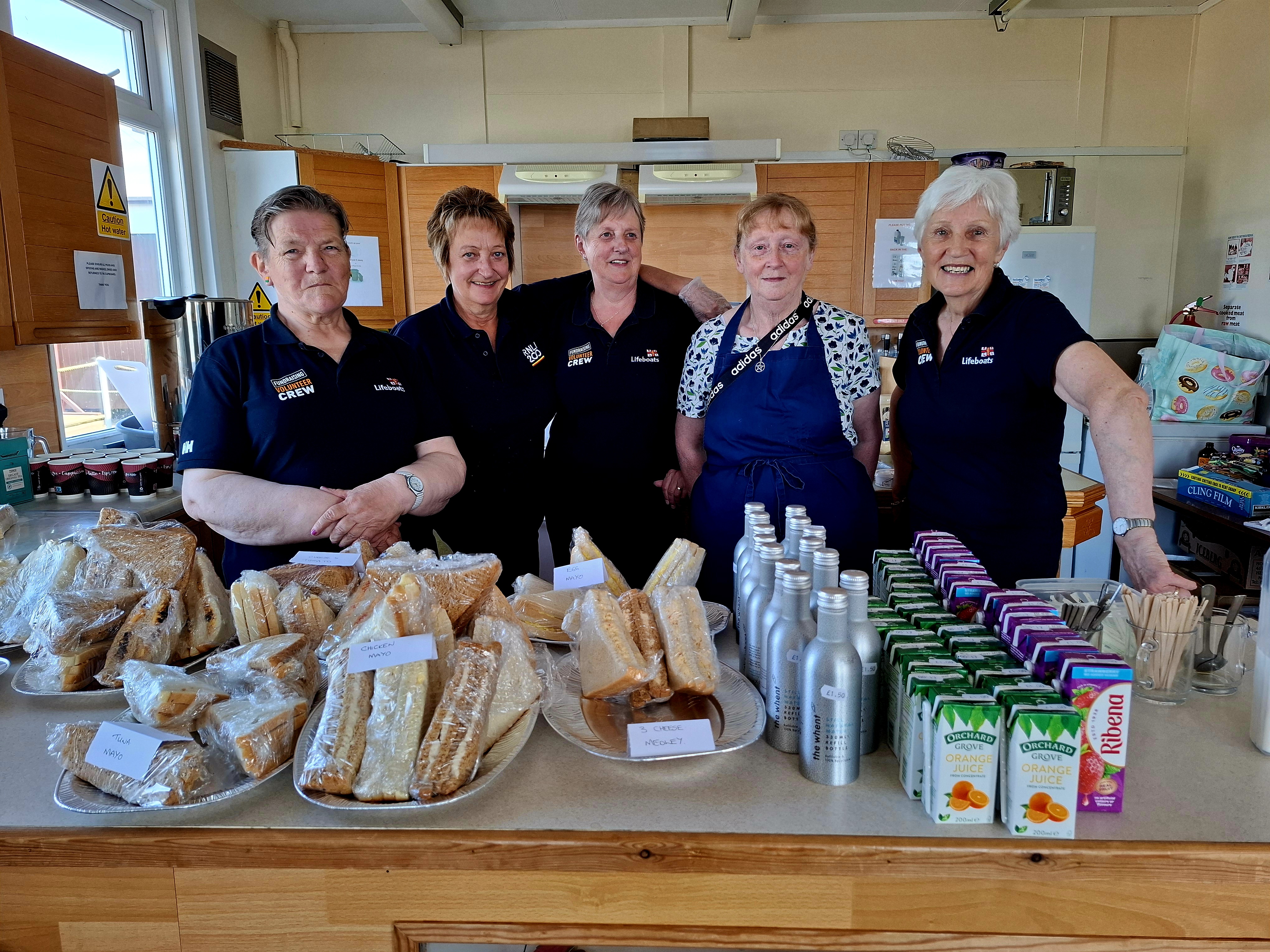 Five fundraising women by food counter with an array of sandwiches and juice.