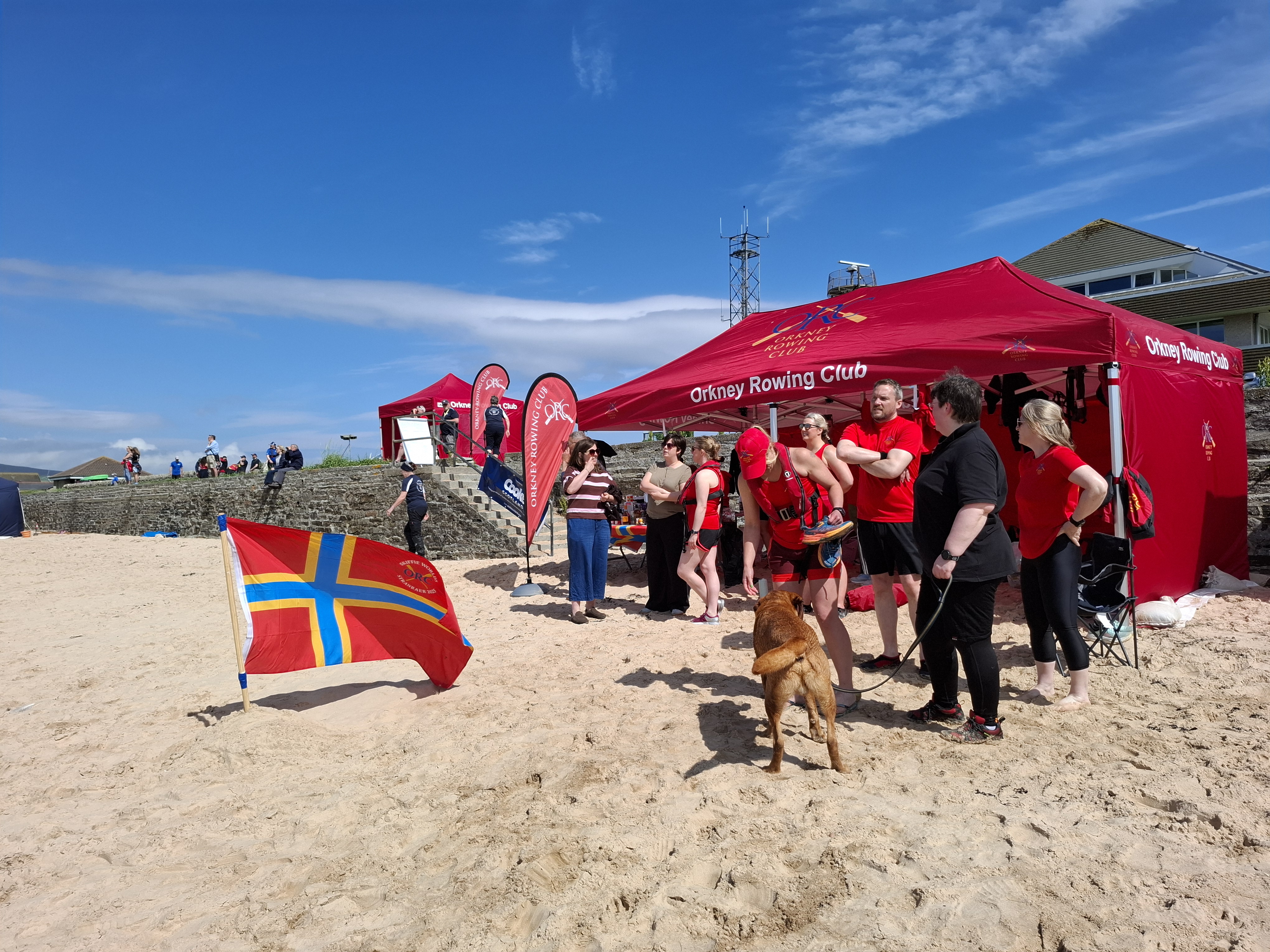 Red gazebo, Orkney flag and people on Scapa beach, with blue skies above.