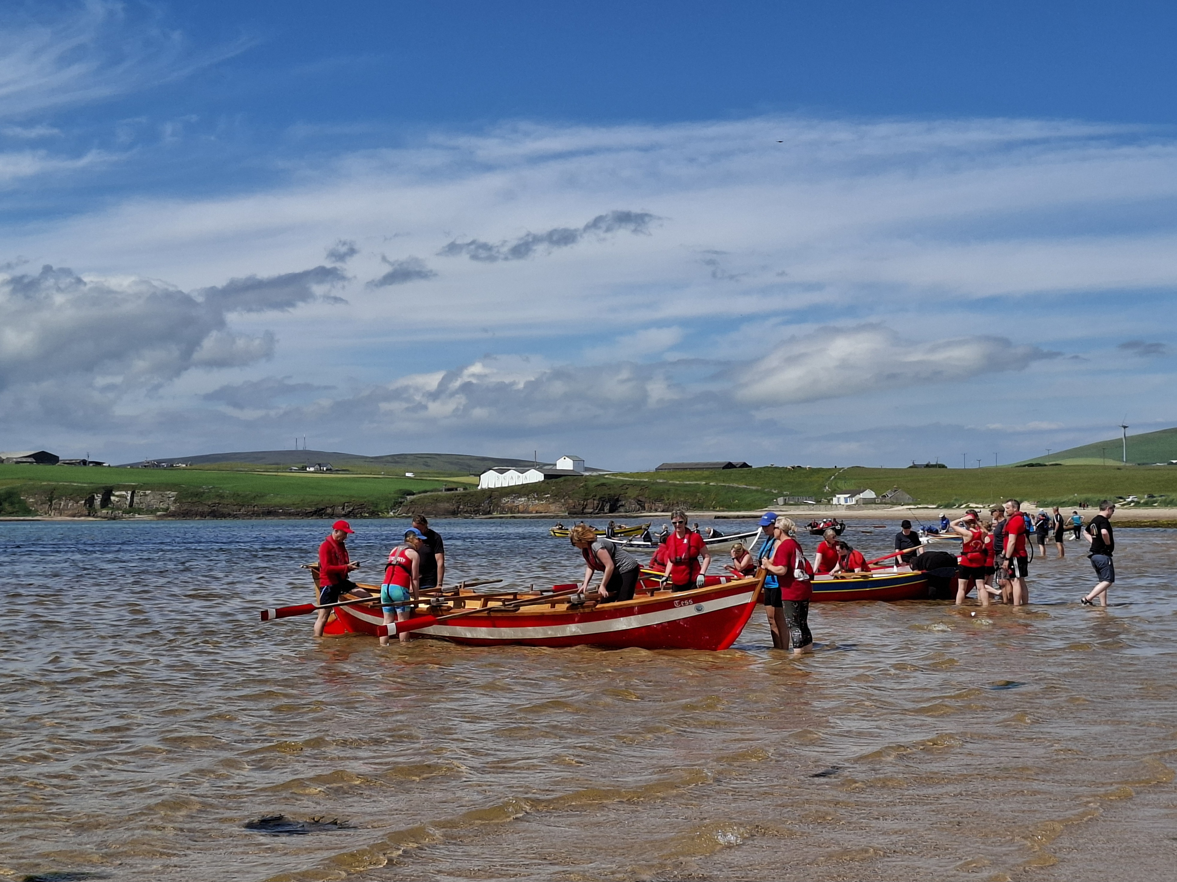 People getting skiffs ready to go out in the bay at Scapa beach, under blue skies with Scapa Distillery in the background