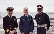 Three men are standing by the coast, one is in a Minehead RNLI jacket and two in ceremonial dress