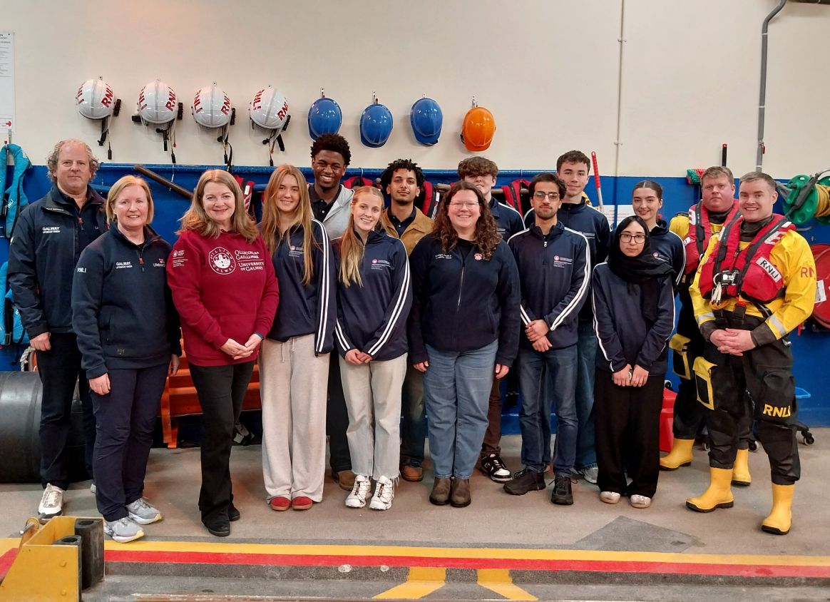 Line up of eleven visitors standing inside the boat house with two crew on left and two crew in dry suits on the right. Helmets, lifejackets and other boat house equipment visible hanging on the wall behind.