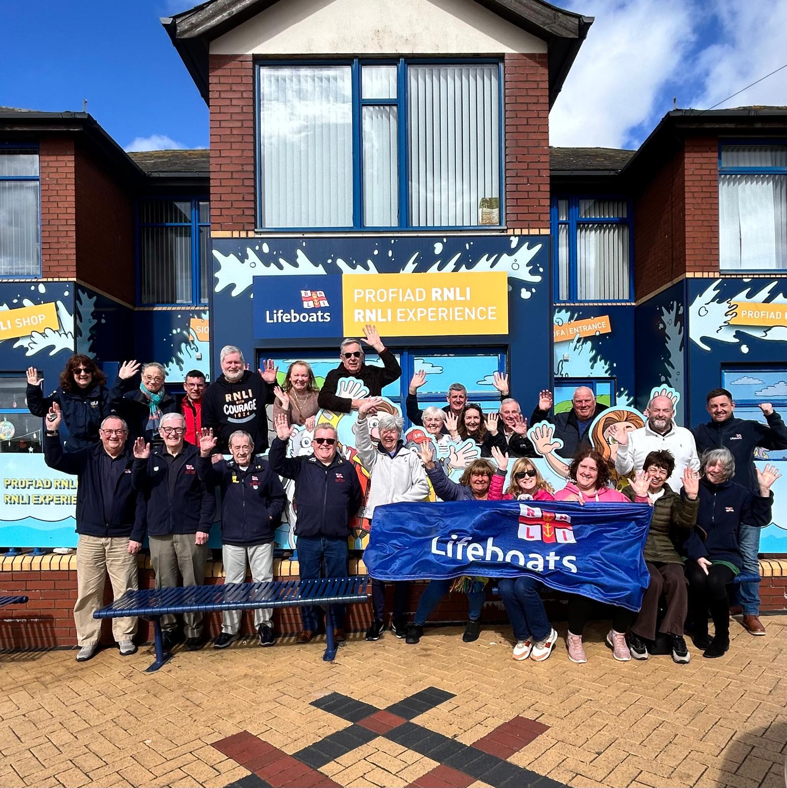 A group of smiling volunteers waving outside the RNLI shop and experience centre at Barry Island