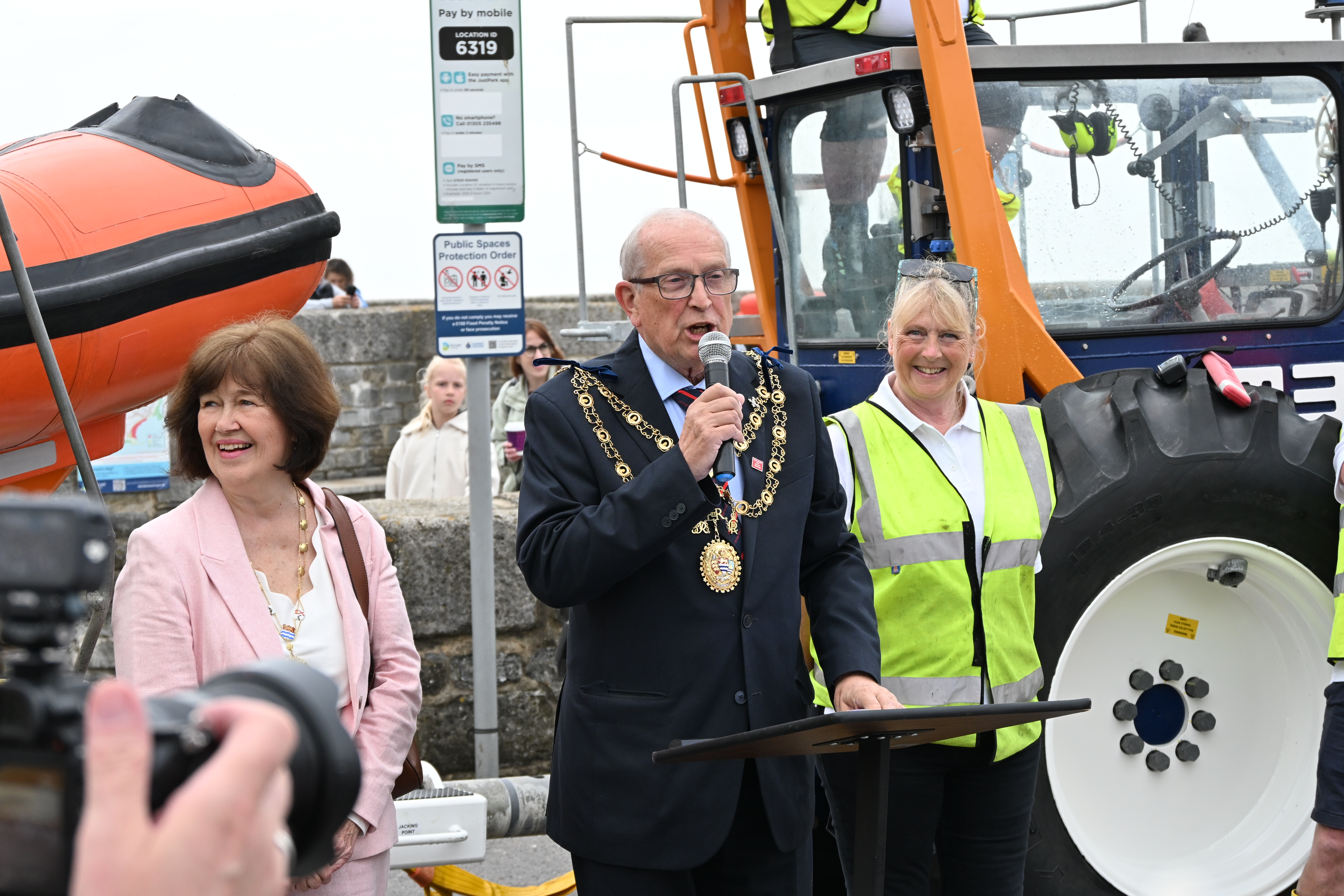 Man in black with microphone flanked by two other people in front of tractor