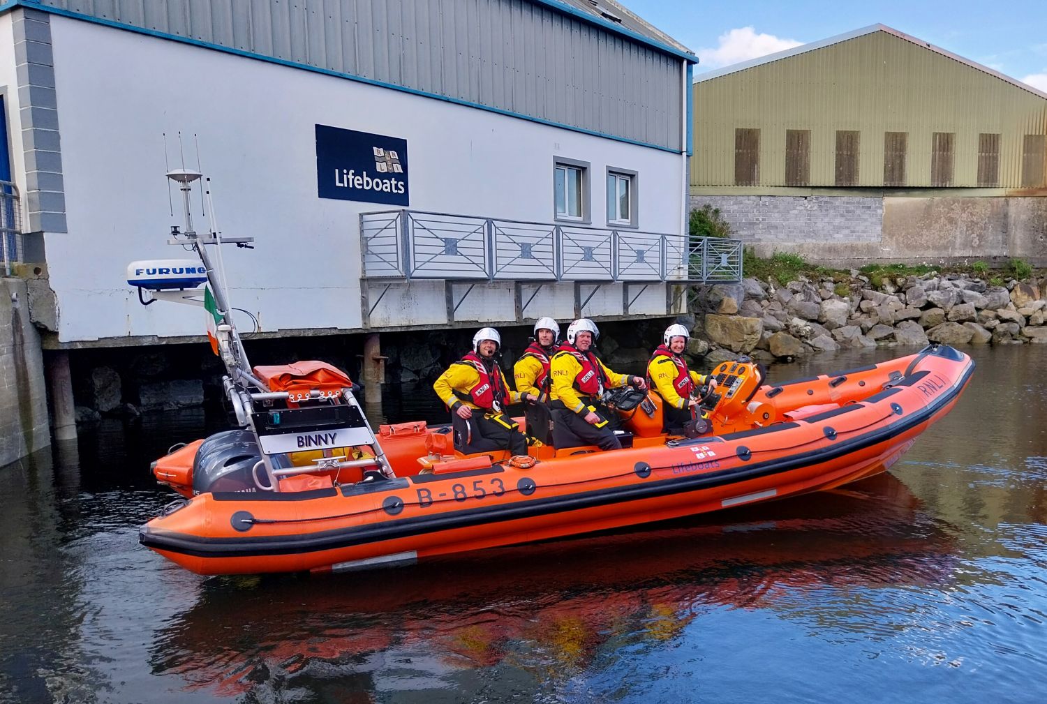 Four crew on the inshore lifeboat just after it launched. The lifeboat station is visible in the background. The crew are all facing the camera for the photo.