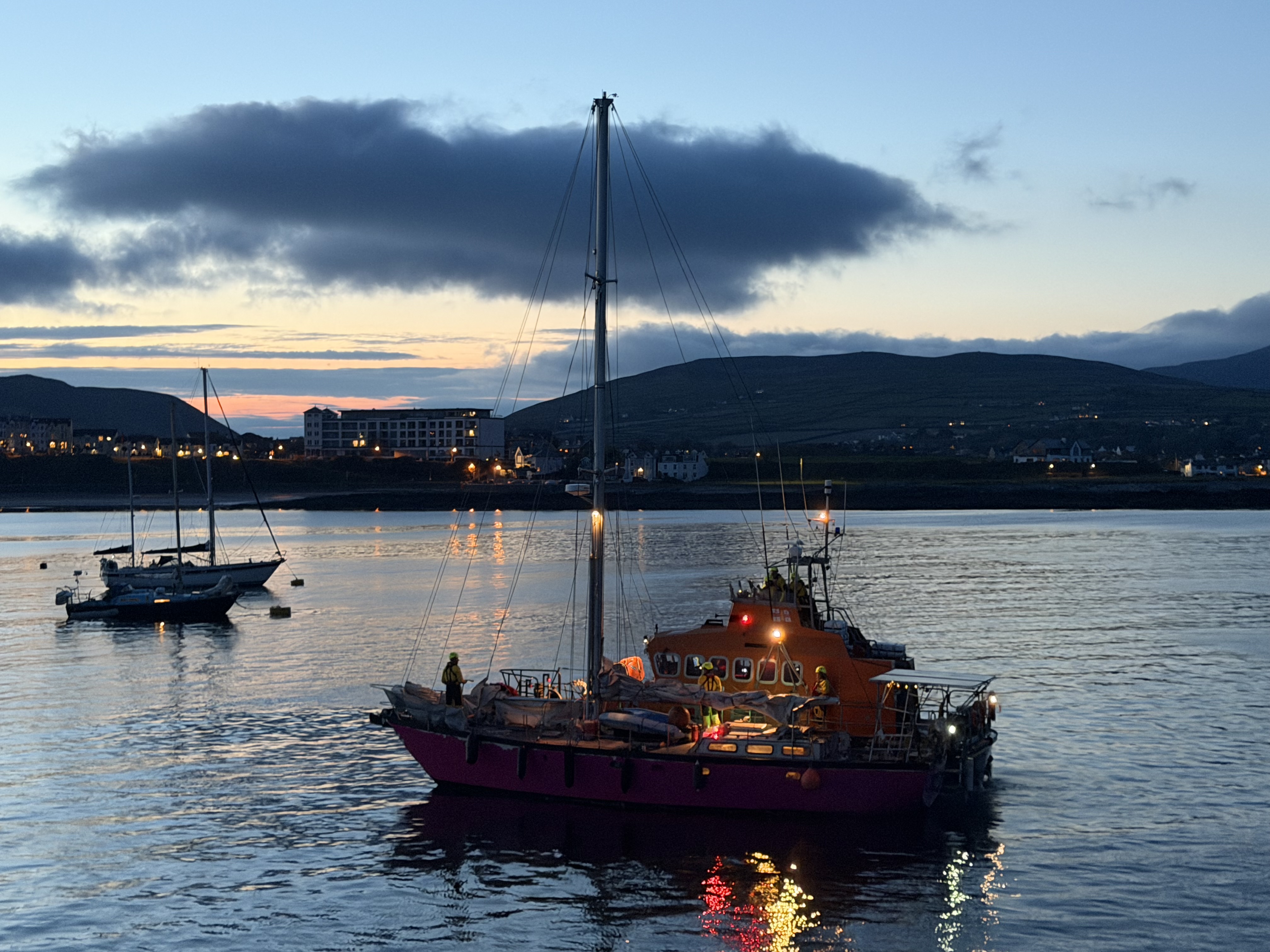 A photo taken at twilight of Port St Mary Lifeboat towing a yacht after its gearbox had failed. Lights on the lifeboat reflect off the sea. There are hills in the background.
