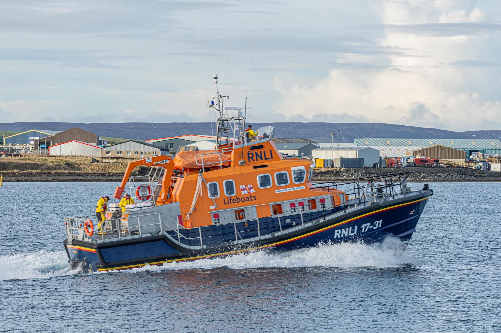 Orange Severn Class All-weather Lifeboat departing from Kirkwall Harbour. In the background are the commercial buildings of Hatston Industrial Estate.
