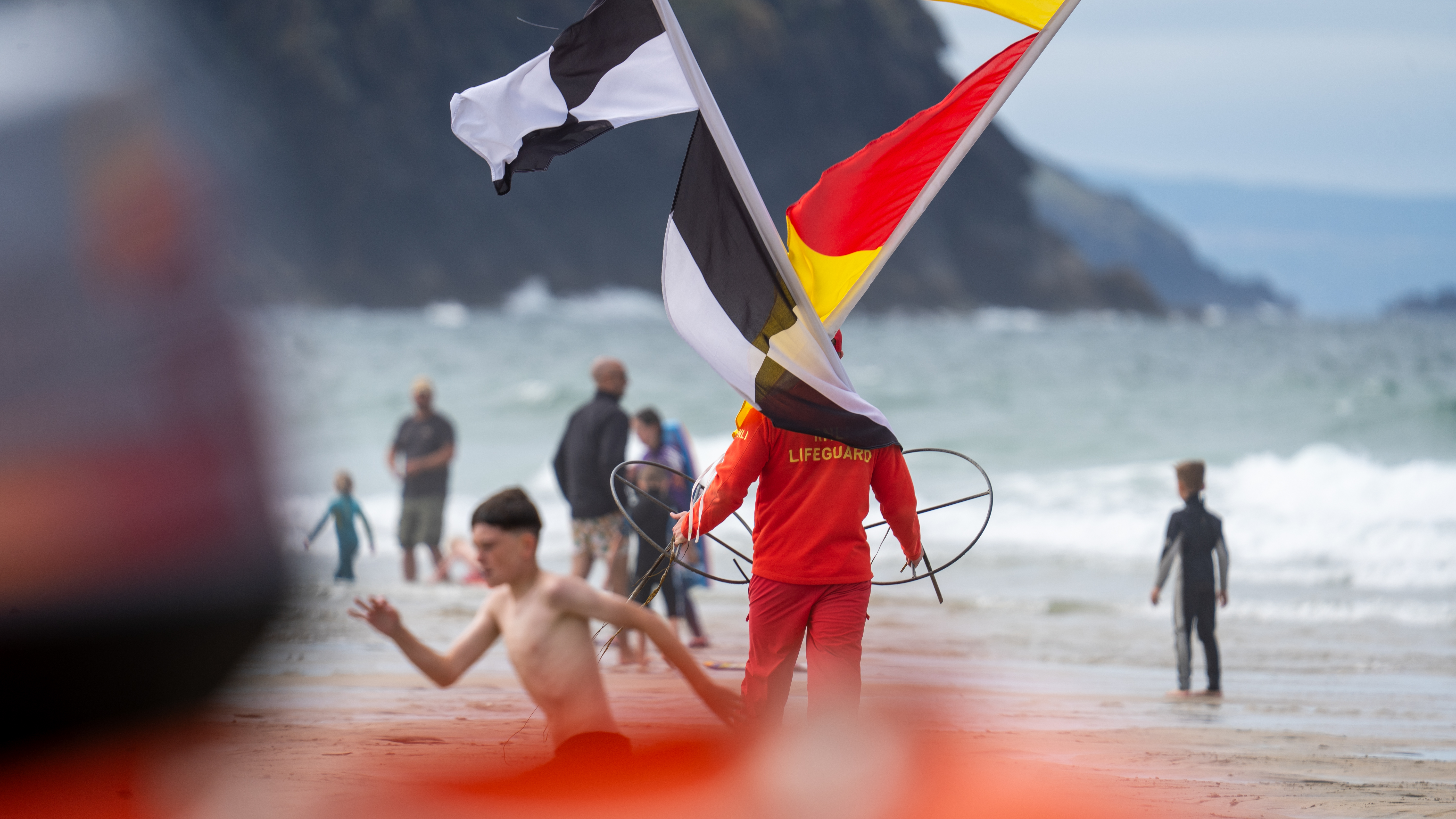 An RNLI lifeguard stood on the beach carrying a black and white flag and a red and yellow flag