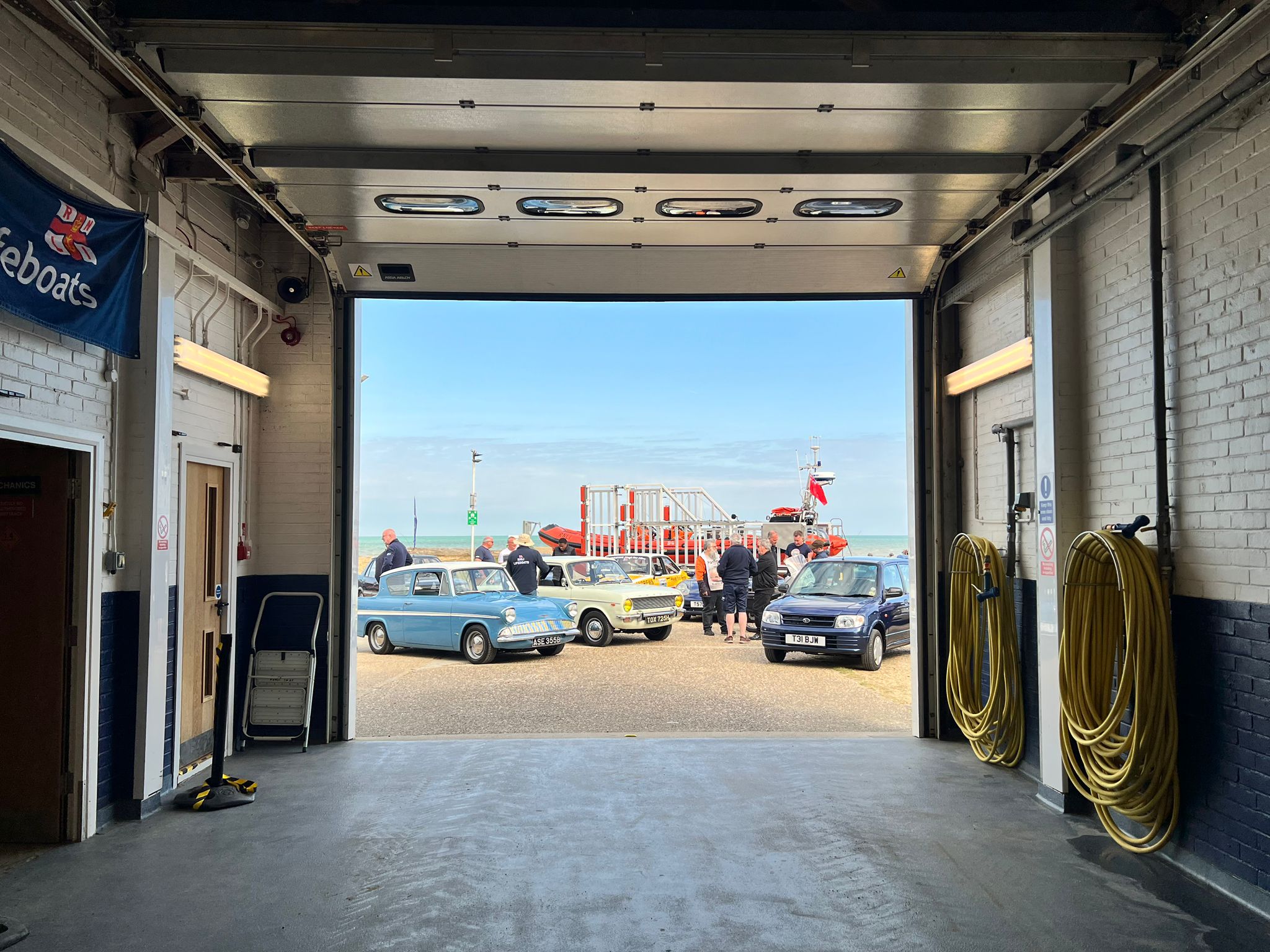 Several old cars infront of the orange lifeboat with the picture being taken from inside the boathouse.