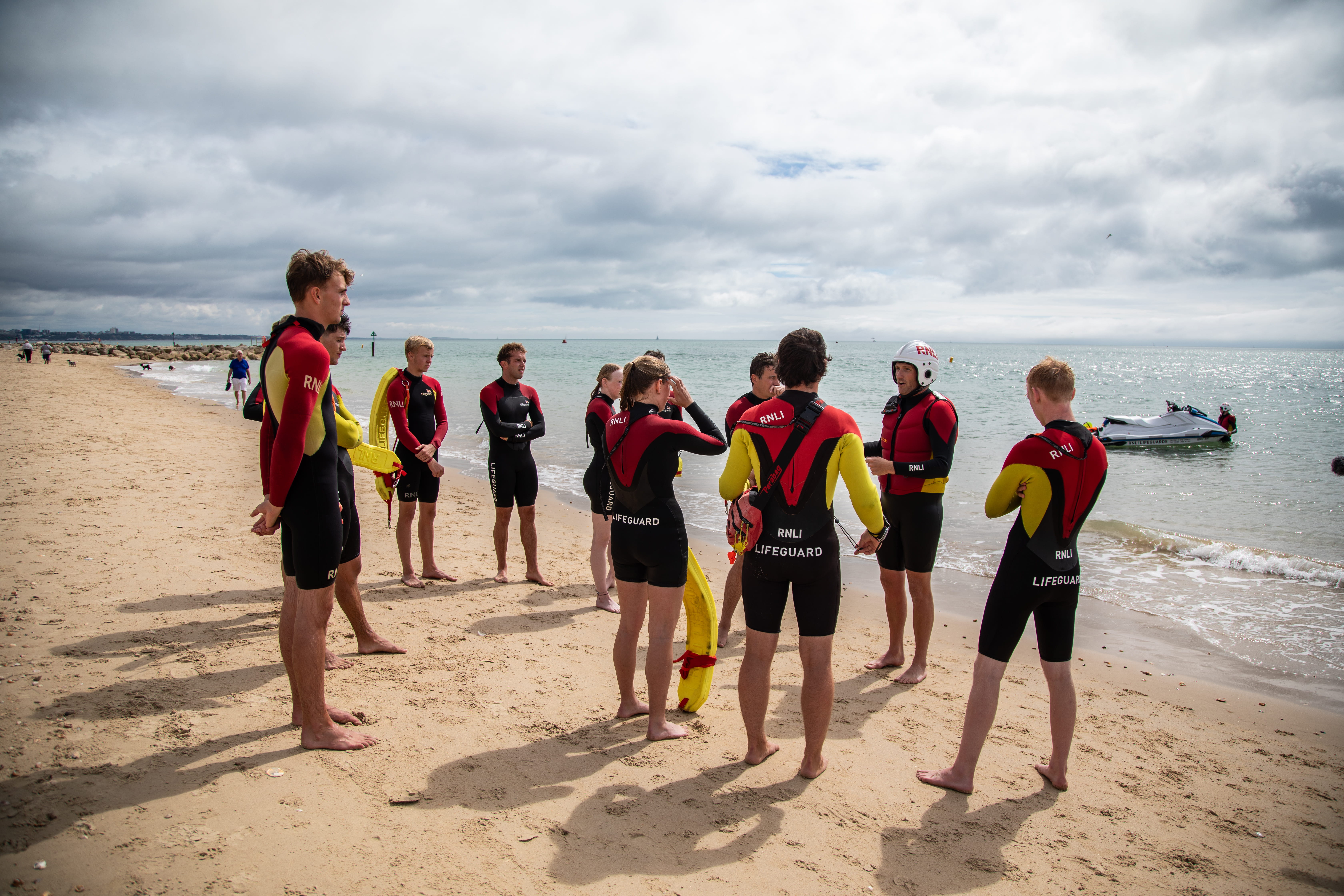 A group of lifeguards stand and listen to an instructor on the beach during a training session.