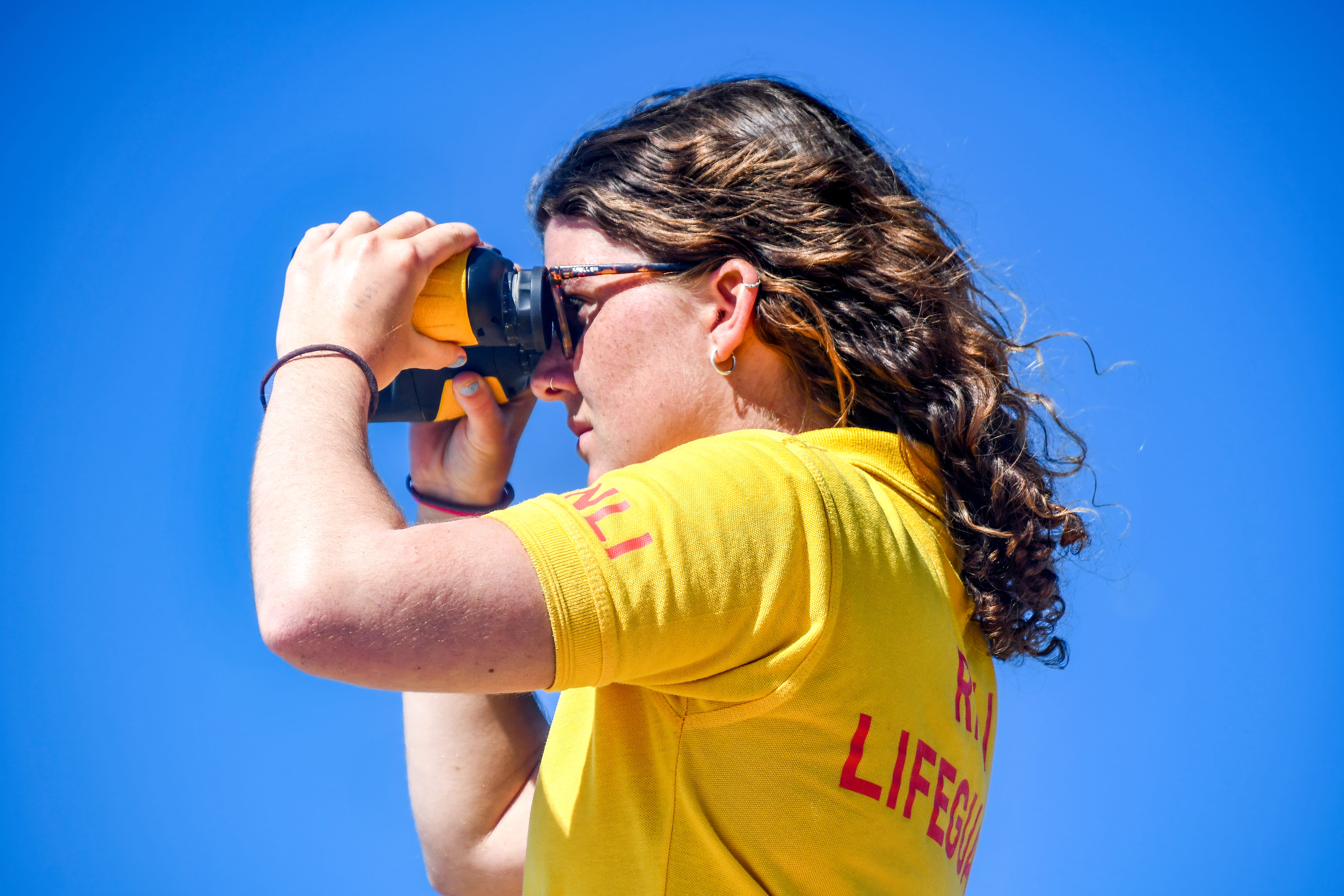 A female lifeguard keeps watch over beachgoers through binoculars on a sunny day.