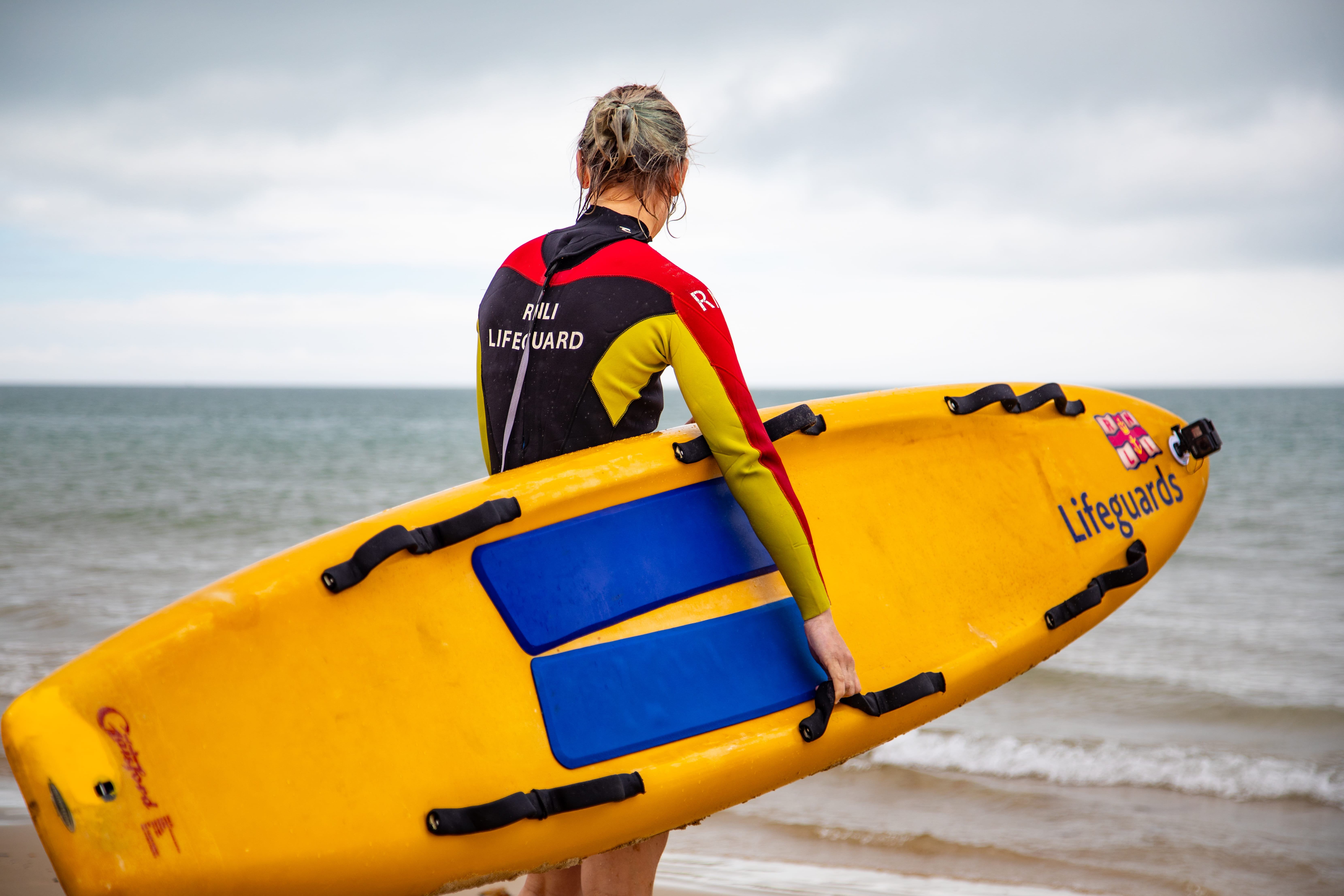 A female lifeguard stands looking towards the sea while carrying an RNLI rescue board under her right arm. Conditions are cloudy and overcast.