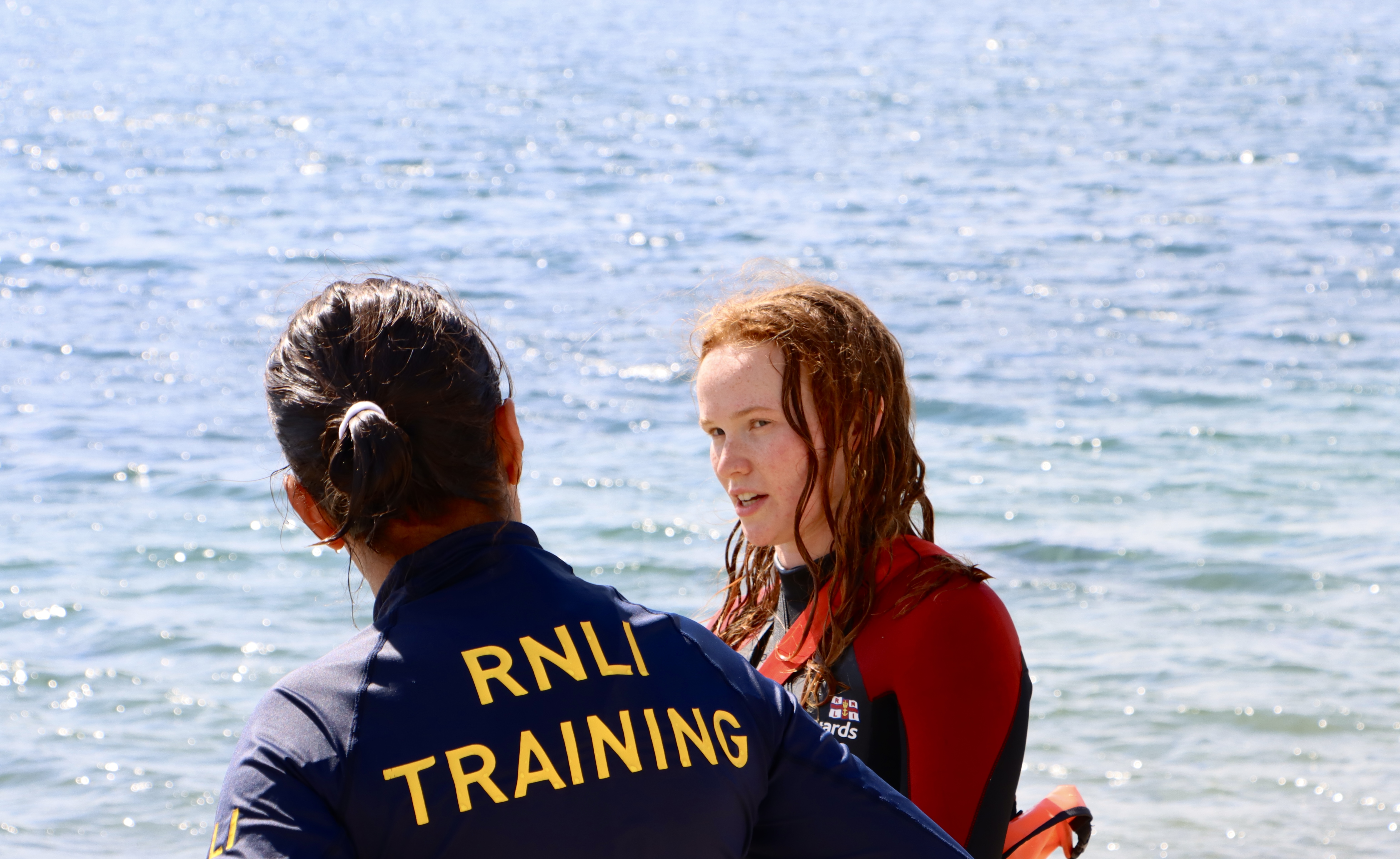 A female lifeguard receives instruction from a female RNLI training advisor by the edge of the sea.