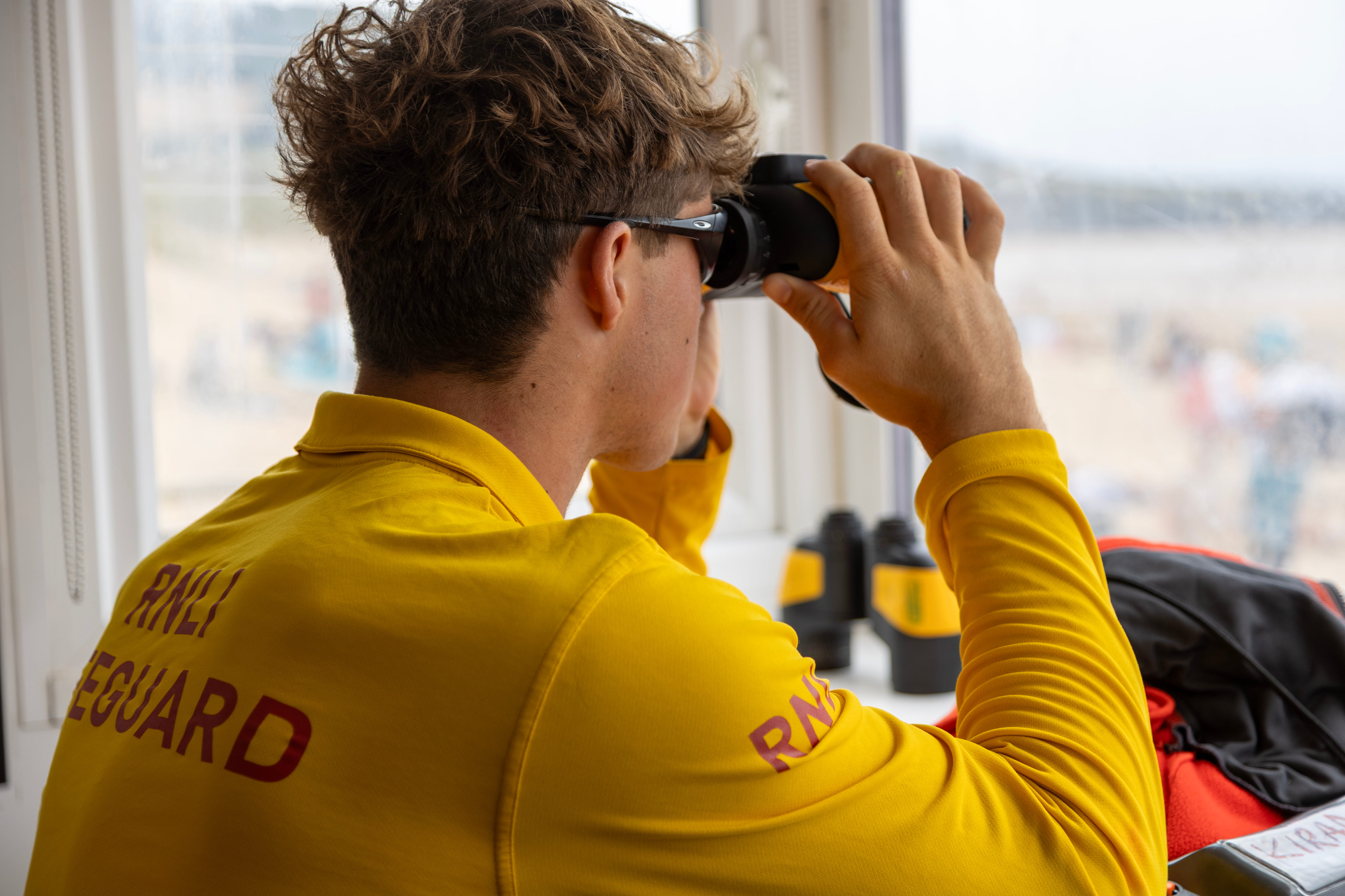 A male RNLI lifeguard keeps watch over beachgoers through binoculars from within a lifeguard beach unit.