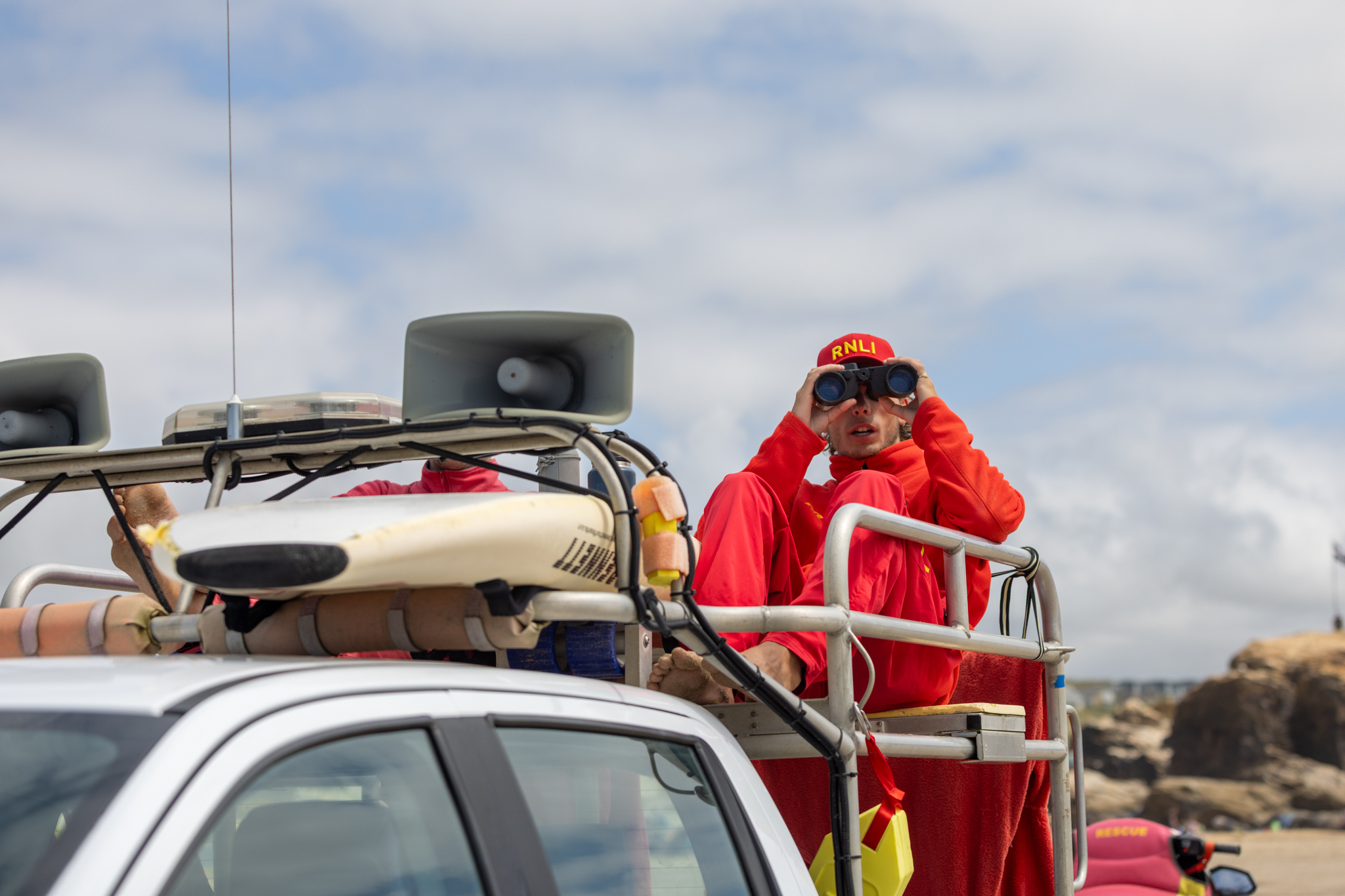 A male lifeguard keeps watch over beachgoers from atop an RNLI rescue vehicle.