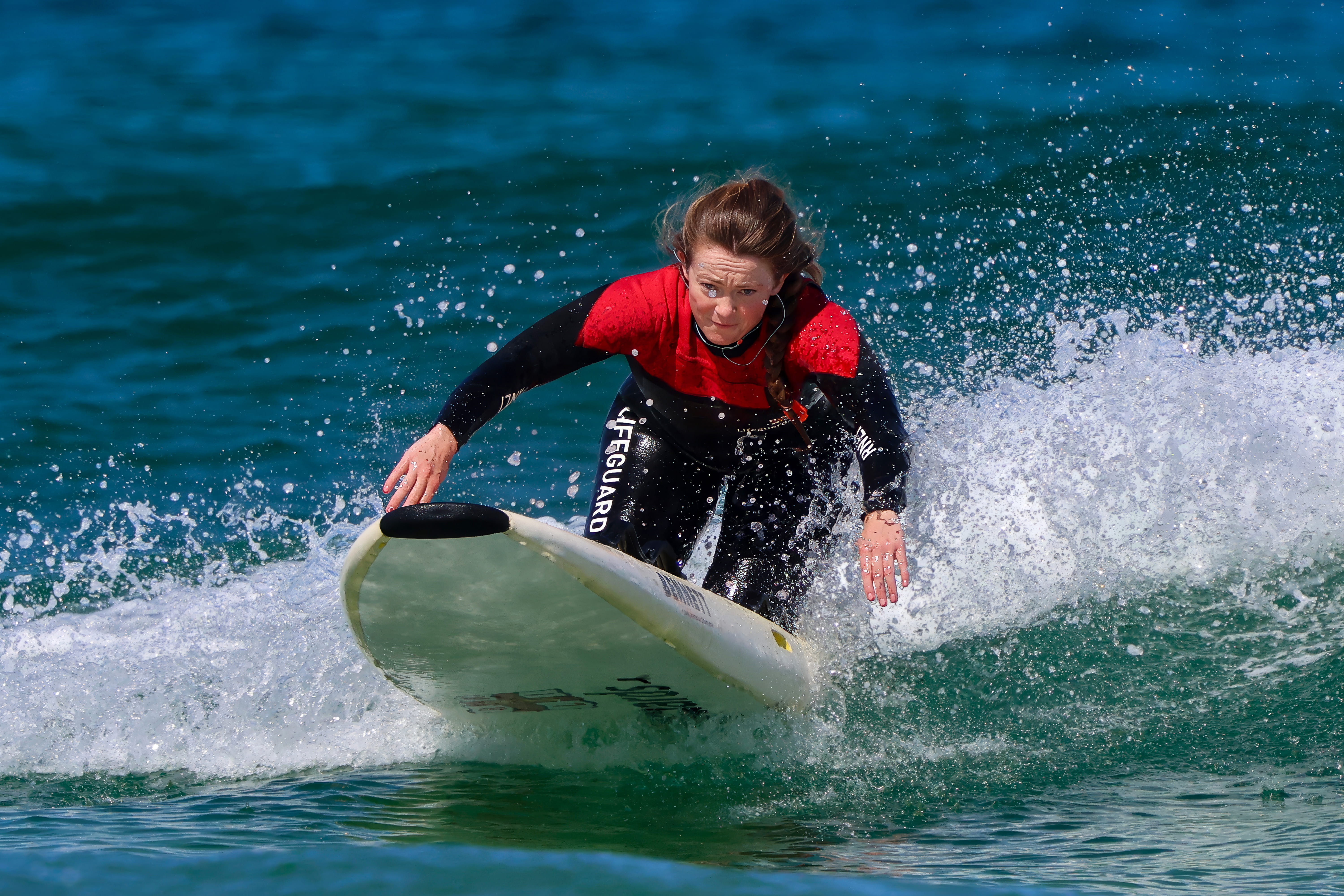 A female lifeguard progresses along the surf atop an RNLI rescue board. The lifeguard wears a wetsuit and conditions are bright and sunny.
