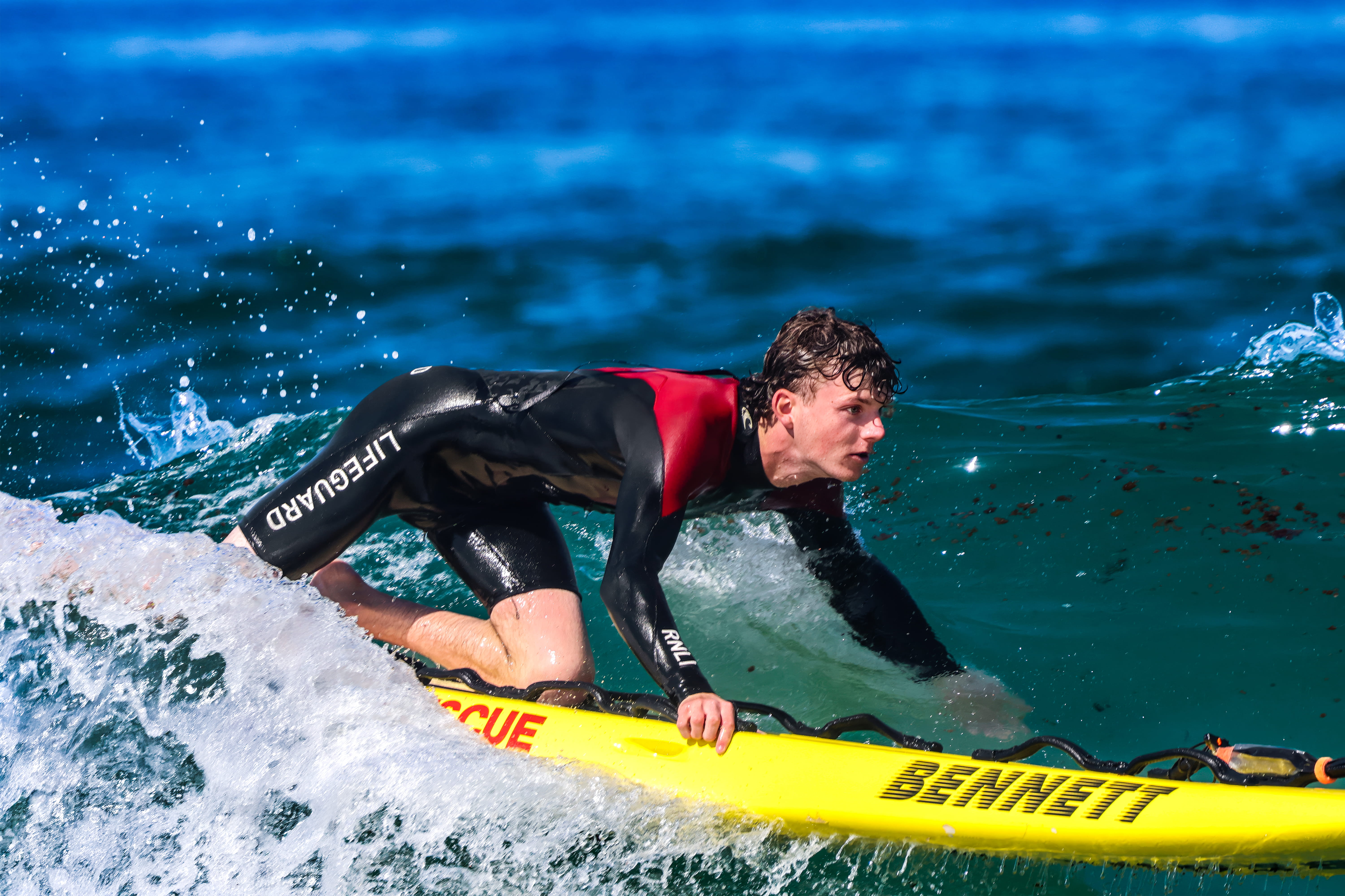 A male lifeguard is crouched atop of an RNLI surfboard as he rides a wave in the sea. The lifeguard wears a wetsuit and the weather is bright and sunny.