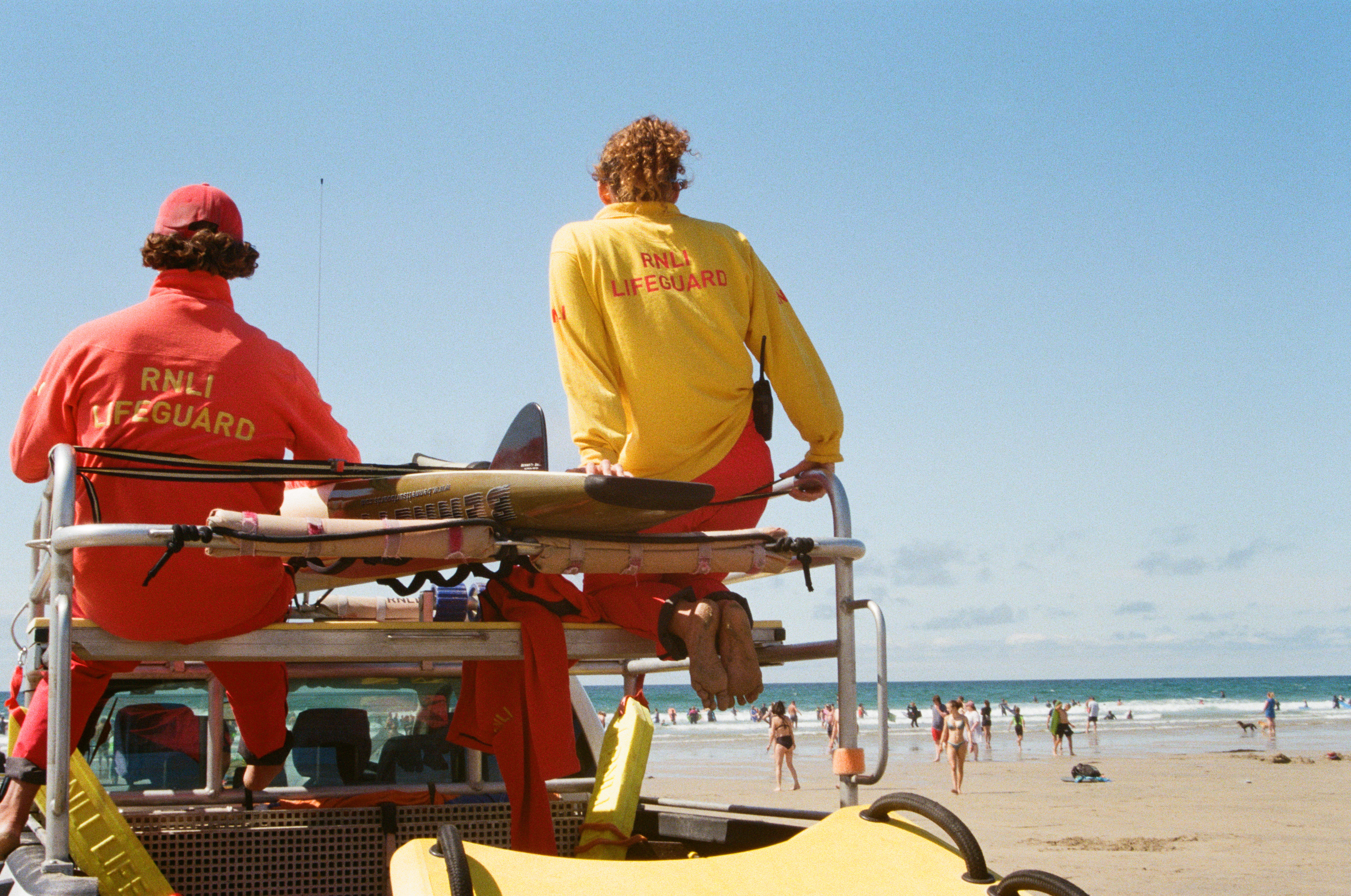 A male and a female lifeguard sit atop an RNLI rescue vehicle as they keep watch on beachgoers on a nearby beach. The weather is bright and sunny. The photographer is positioned behind the lifeguards.
