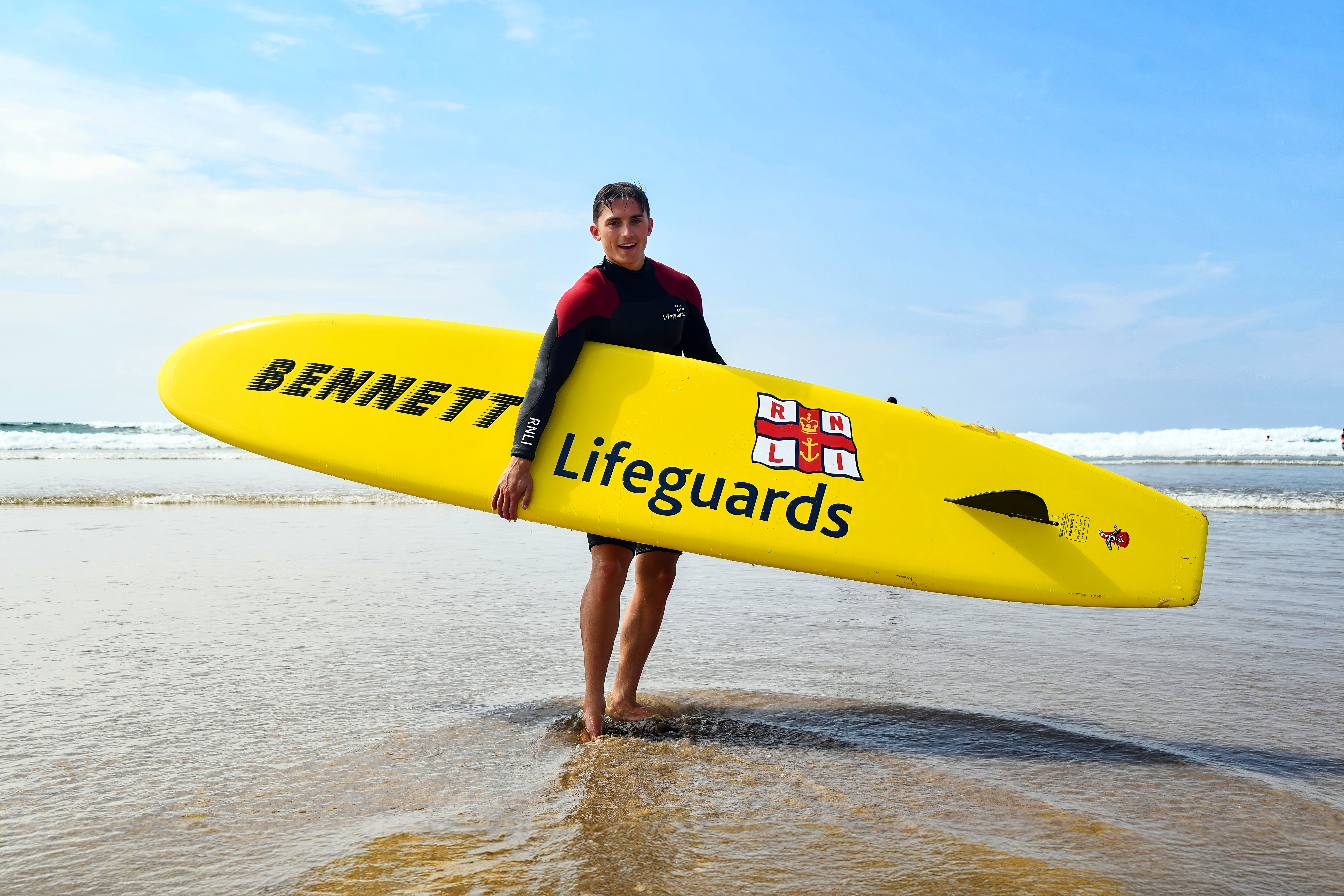 A male lifeguard faces the camera as he stands on a beach with an RNLI rescue board tucked under his right arm. The logo for the RNLI and text with 'Bennett' and 'Lifeguards' can be seen on the underside of the board. Sea water partly submerges the lifeguard's feet.