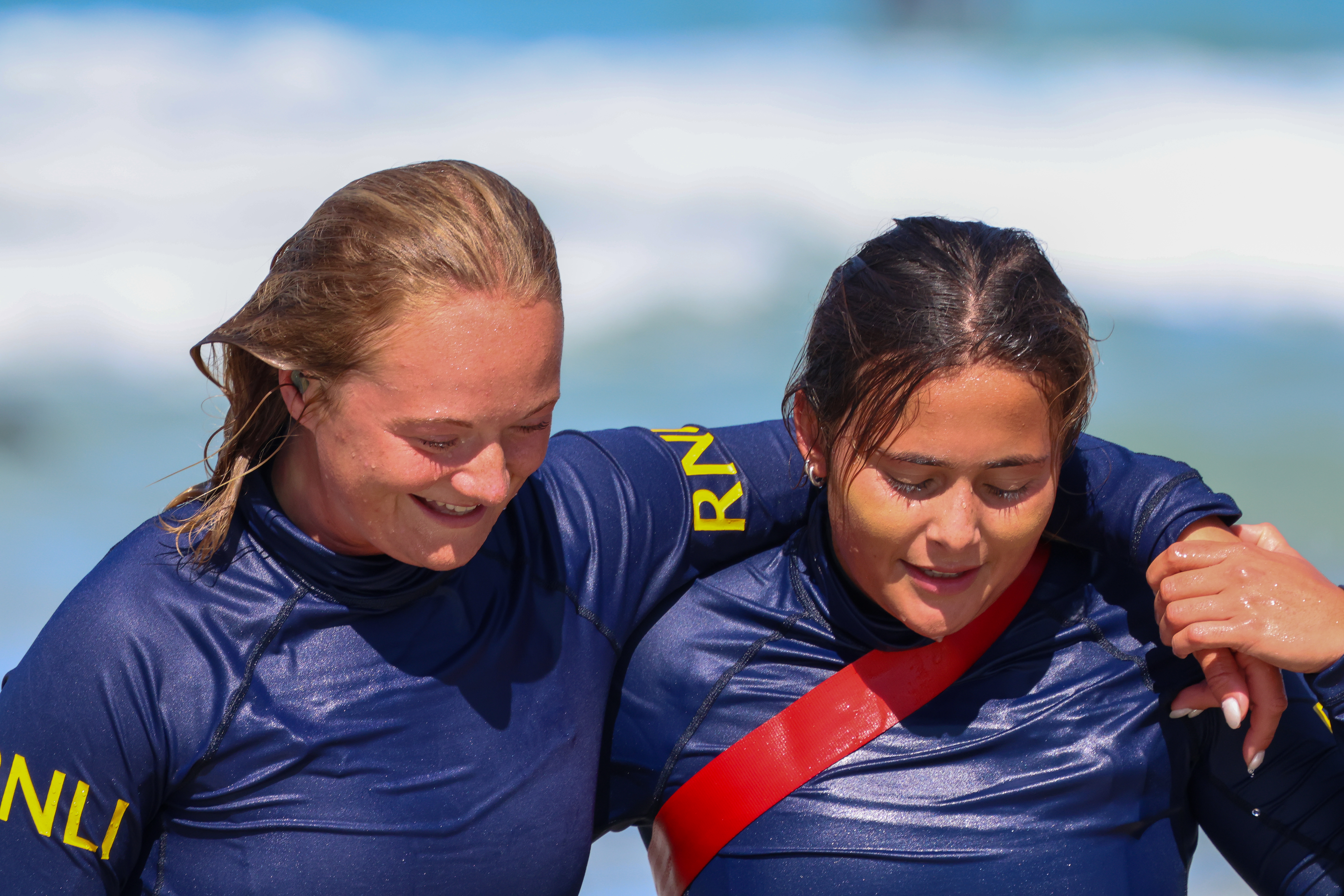 Two female lifeguards practice rescue techniques as part of their pre-season training. One lifeguard supports the other as they walk together. Both lifeguards are smiling.