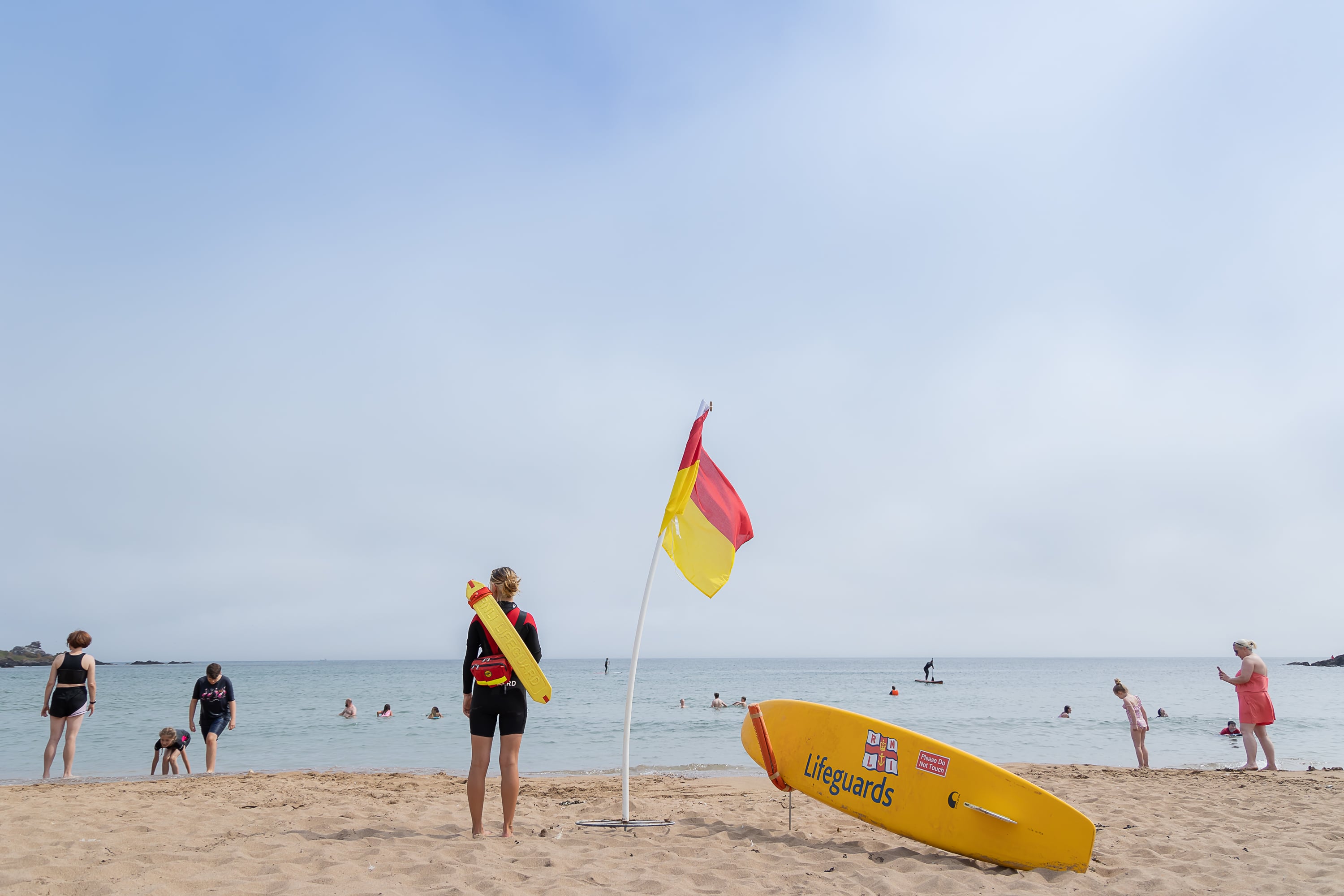 A female lifeguard stands on watch upon a beach while members of the public enjoy a sunny day at the coast.