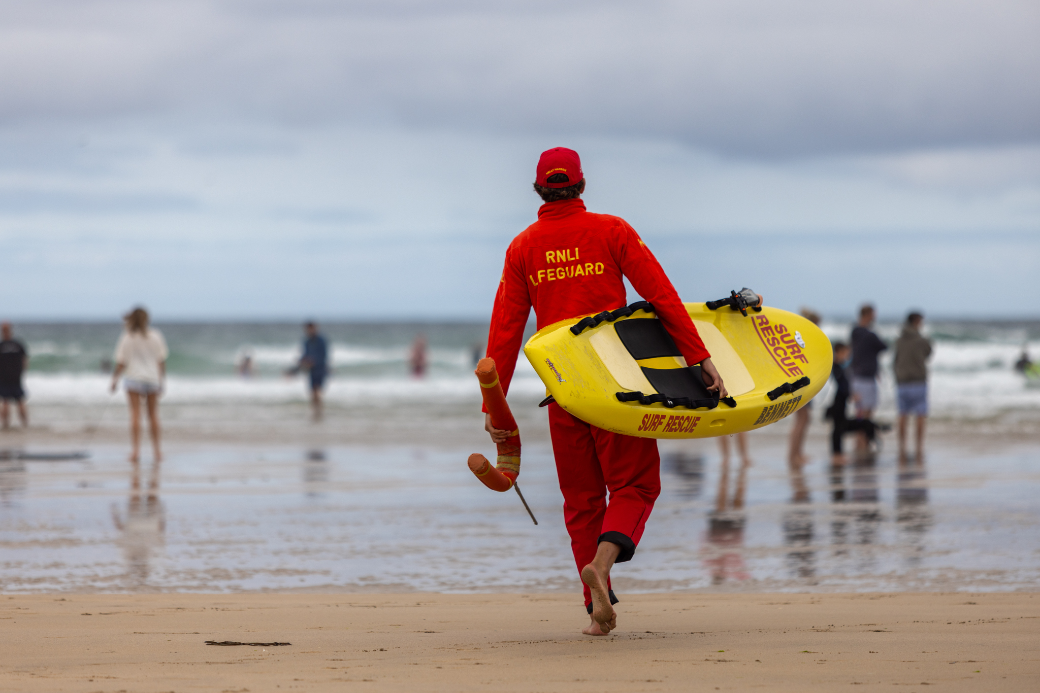 A male lifeguard walks towards the sea while carrying an RNLI rescue board under his right arm and a buoyancy aid in his left hand.