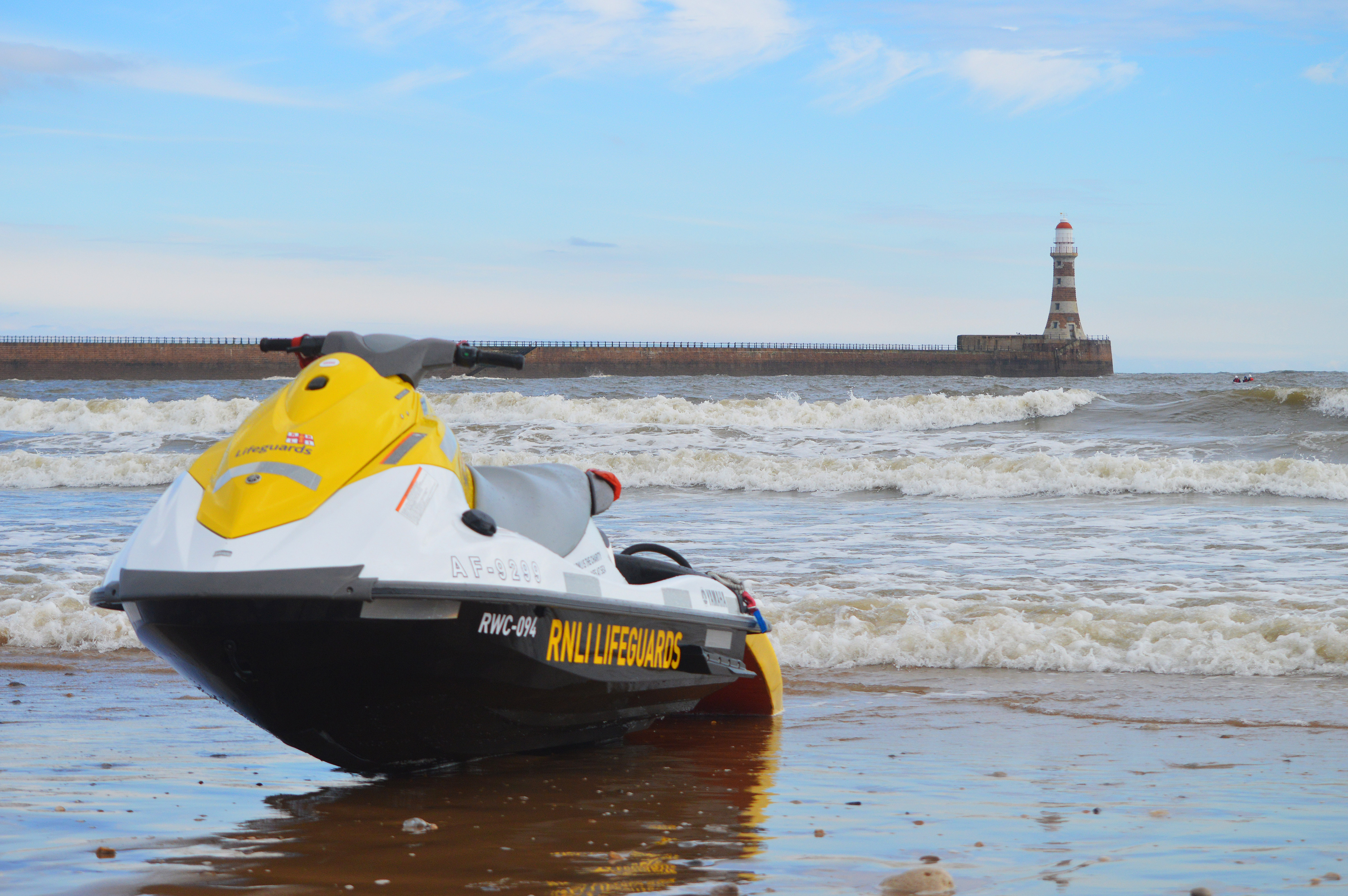 An RNLI rescue craft sits on the beach as the tide comes in from beach. A sea defence and striped lighthouse can be seen in the background.