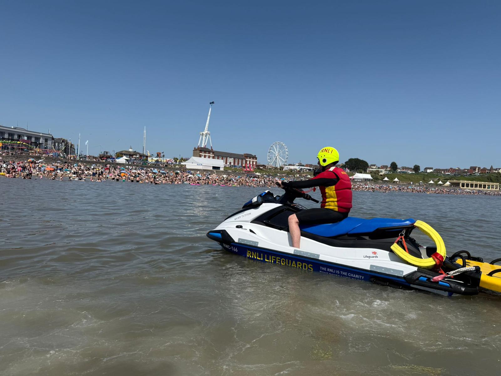 The photo shows a lifeguard on a rescue water craft in the sea looking back at a very busy beach