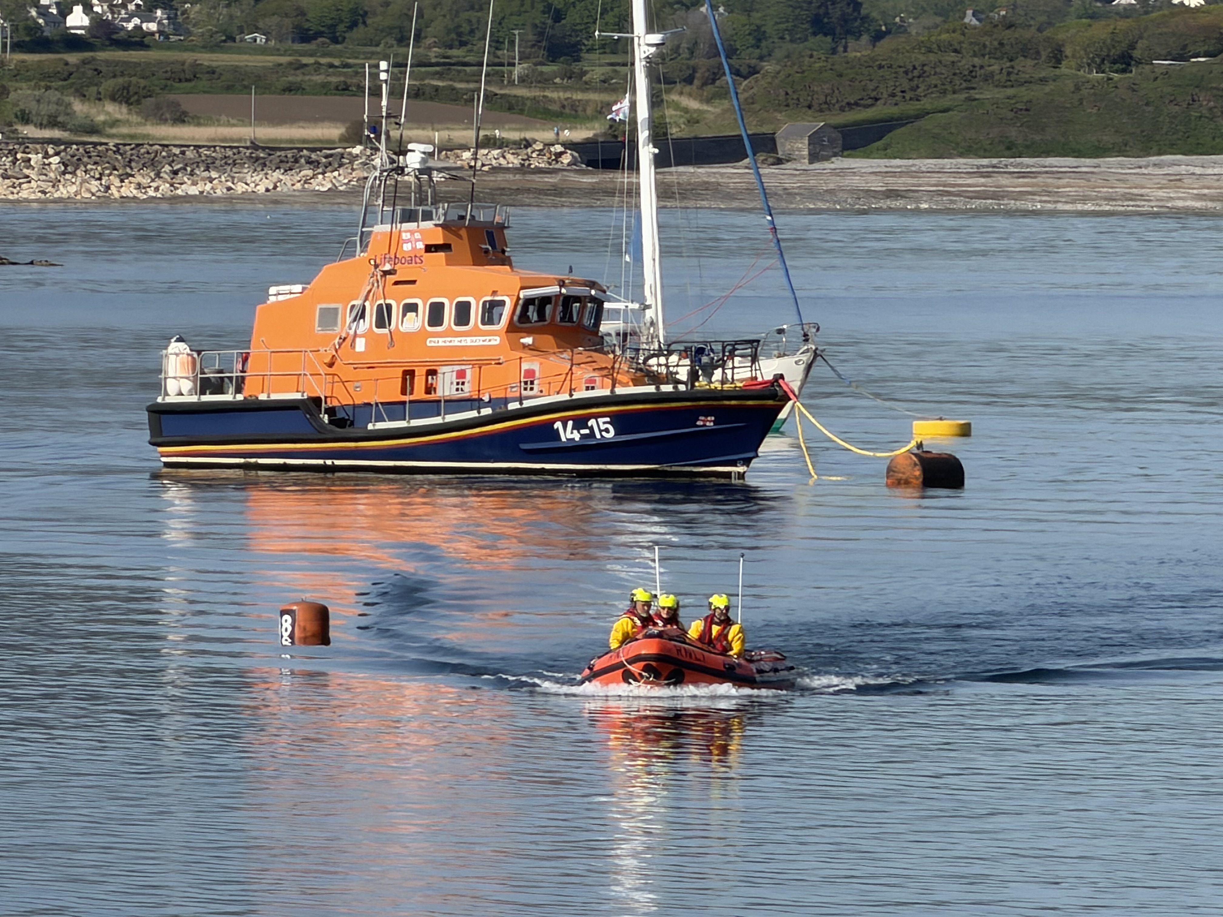 Picture shows the inshore lifeboat with 3 crew members on board. The sea is calm. The bigger Port St Mary Lifeboat is in the background moored up. Hills are in distance. It is a lovely sunny day.