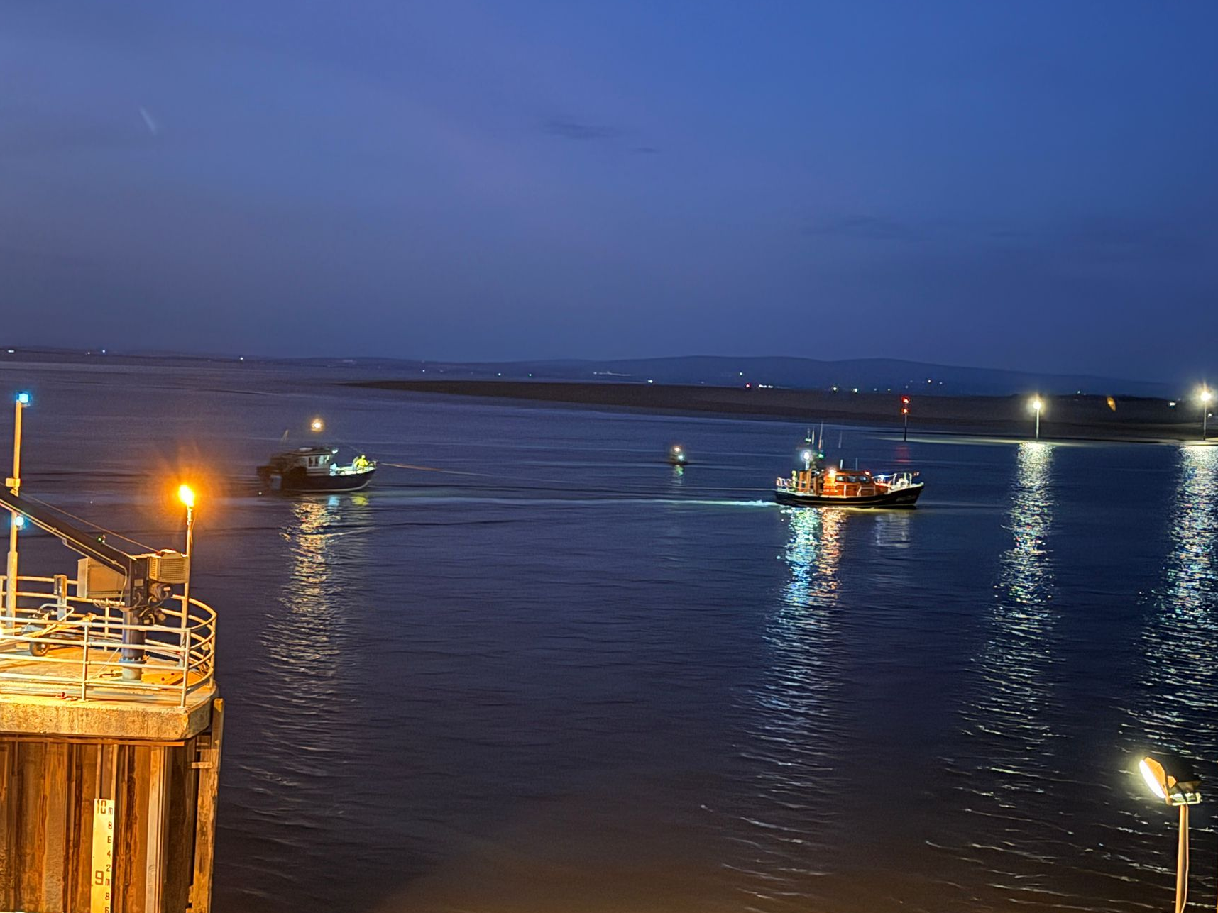 A Shannon class lifeboat towing a small commercial fishing vessel at night. The lights of RNLI Fleetwood's mooring pen are in the foreground.