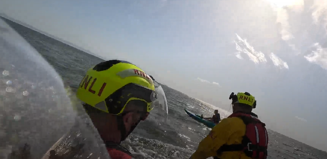View from Barry Dock RNLI's D-class inshore lifeboat as volunteer crew approach a blue and green kayak floating on the sea. The water is calm with a slight swell and bright sunshine reflects off the surface.
