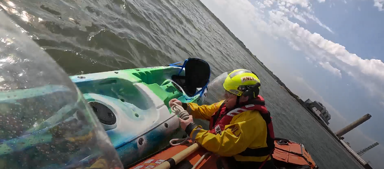 An RNLI crew member wearing yellow protective clothing and a helmet leans from the orange inshore lifeboat to secure a blue and green kayak floating alongside.