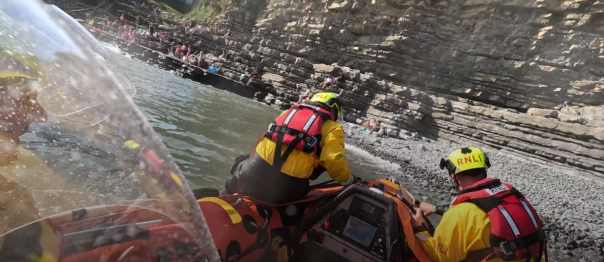 The RNLI inshore lifeboat approaches a rocky beach beneath steep cliffs, where people can be seen watching from the shore.
