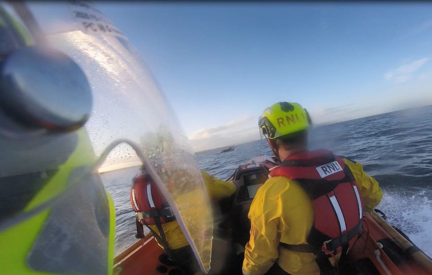 A shot of crew members in the foreground aboard D-Class lifeboat, visible in the distance is a dark coloured vehicle almost fully submerged.