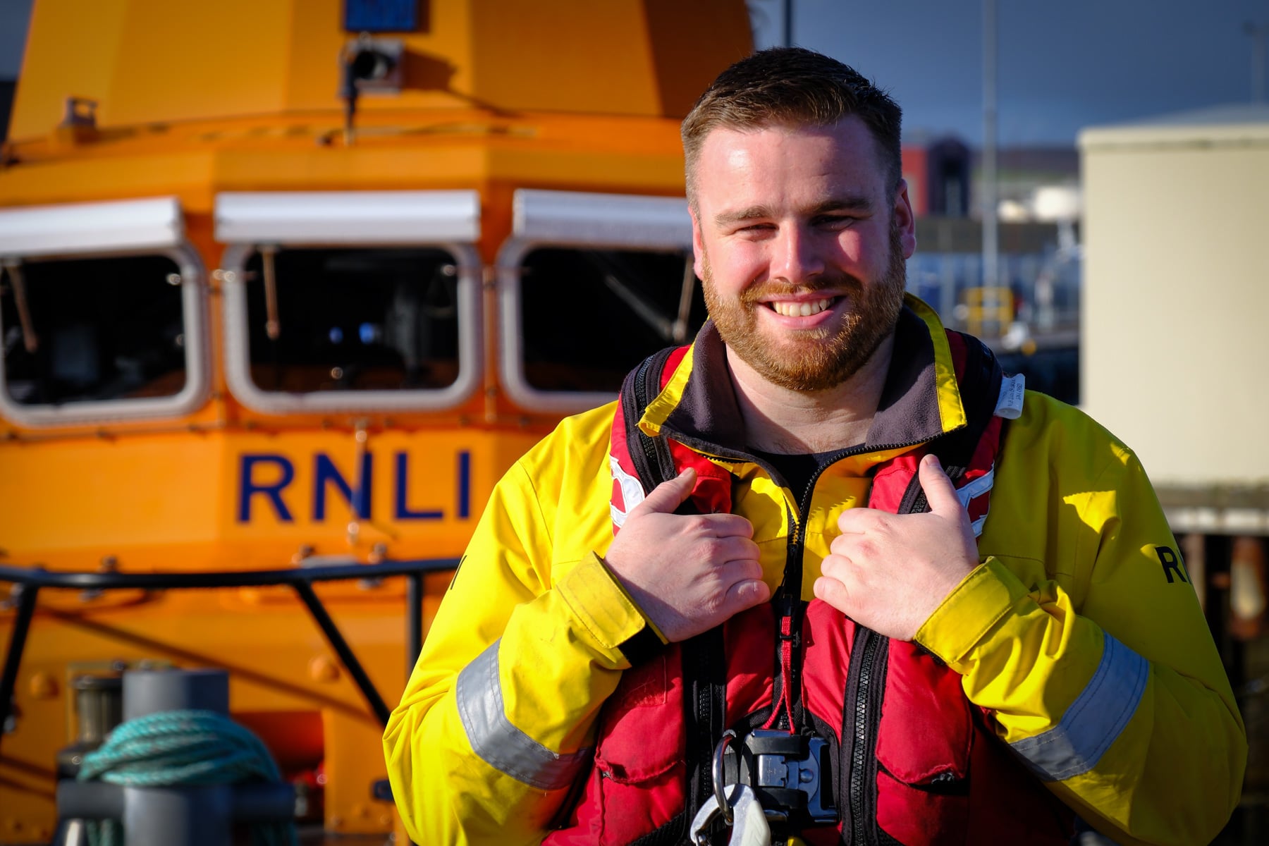 Photograph of Cammy Barron, standing sunlit, in yellow RNLI gear with red lifejacket, looking at camera with the lifeboat behind