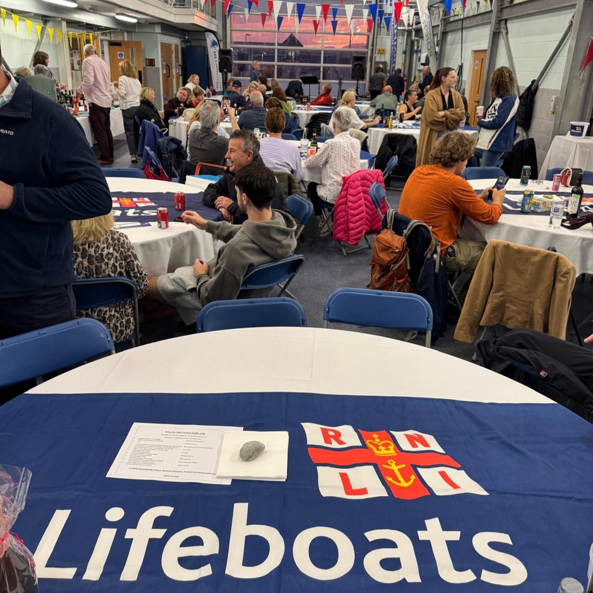 people sitting around tables inside Pwllheli RNLI lifeboat station, and the pink sunset can be seen through the large door in background.